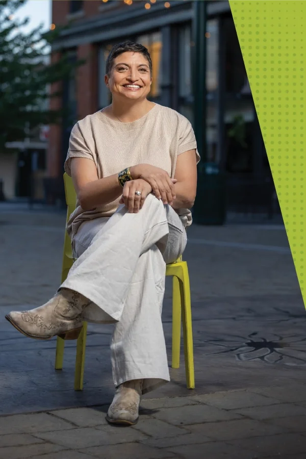 Janelle Carman-Wagner (JCW) sitting on a lime green chair outdoors, smiling, wearing a beige top, white pants, and cowboy boots at first and Main in downtown Walla Walla