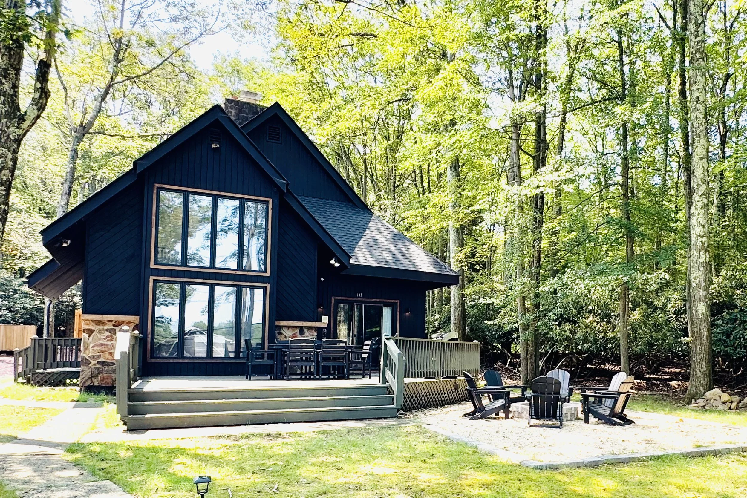 A modern, dark blue, two-story house with large glass windows, a stone pillar, and a backyard patio with outdoor chairs and a fire pit, surrounded by tall green trees.
