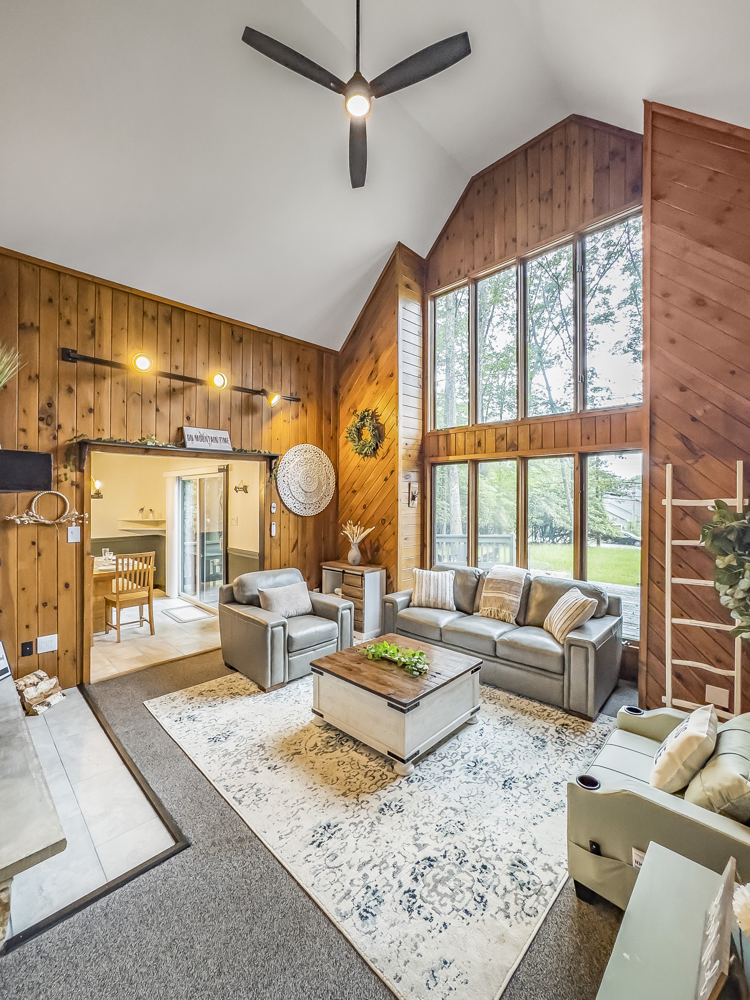 Living room with large wooden wall panels, high ceiling, and big windows showing trees outside. There are two gray sofas, a white wooden coffee table, and a white armchair. Twin sized pull out bed.