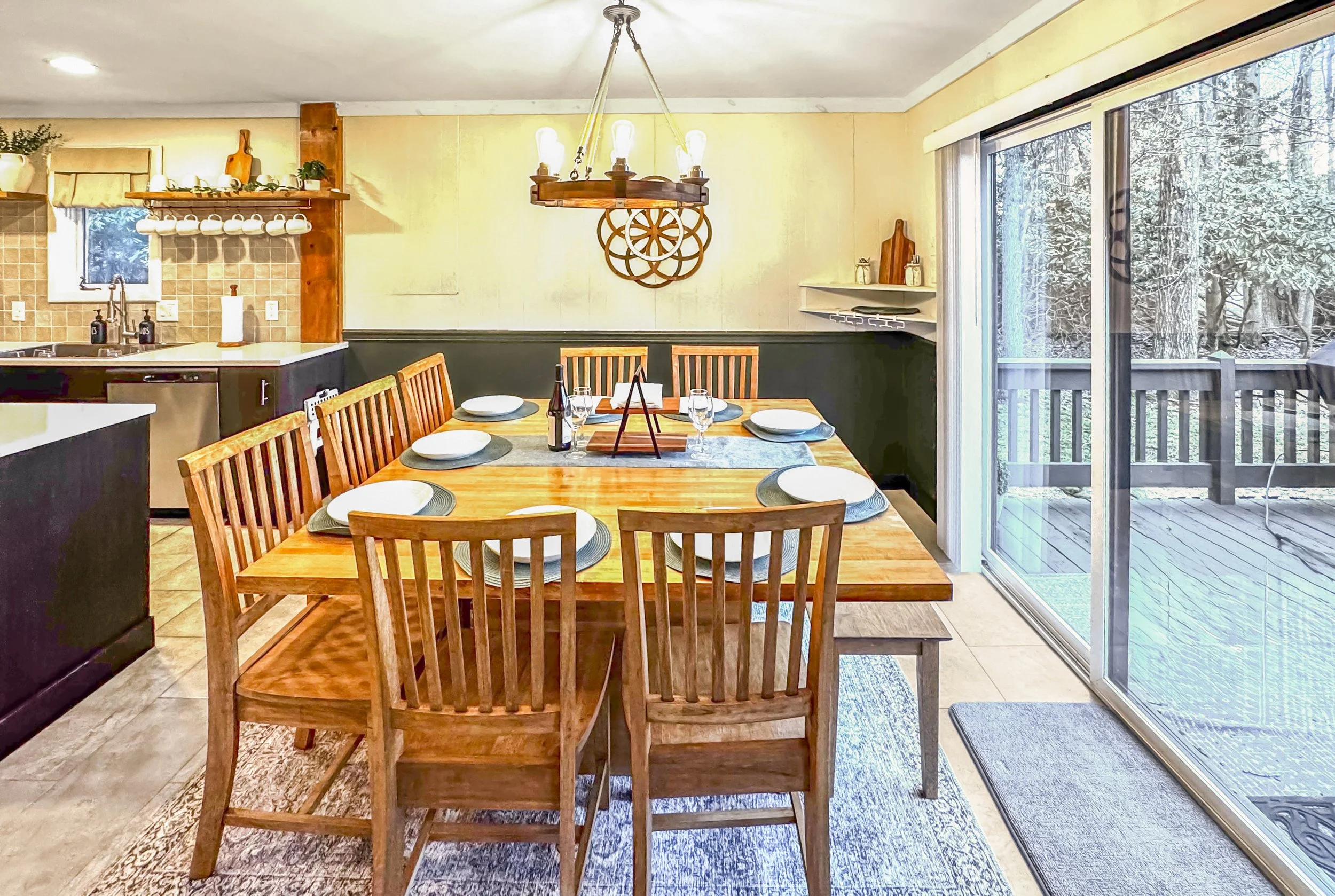 Dining area with a wooden table set for six, with plates, glasses, and napkins, adjacent to large sliding glass doors leading to a deck with a lake view outside.