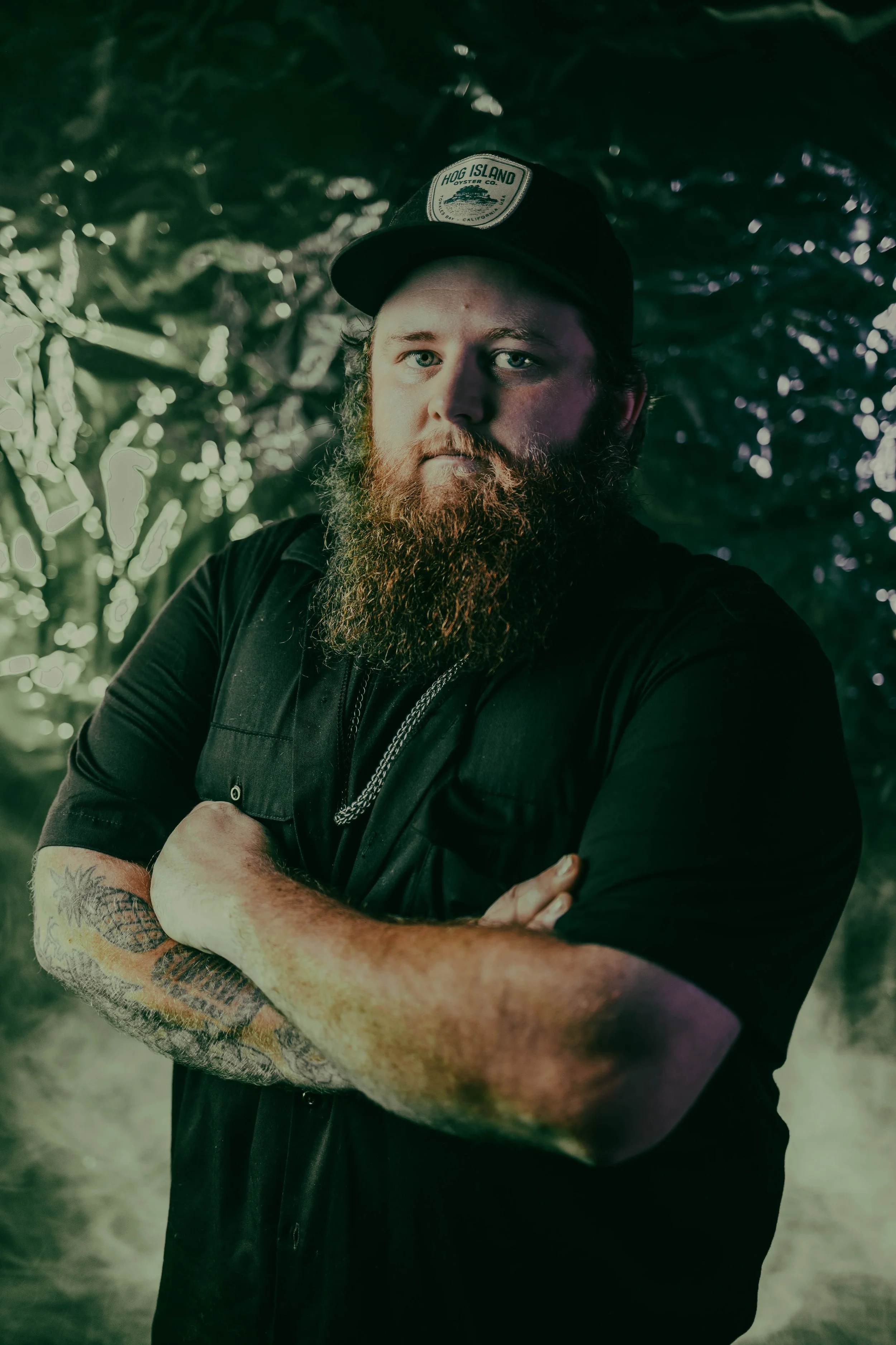 A man with a beard and tattooed arms wearing a black shirt and baseball cap crossing his arms, standing in an outdoor setting with a blurred background of trees and bokeh lights.