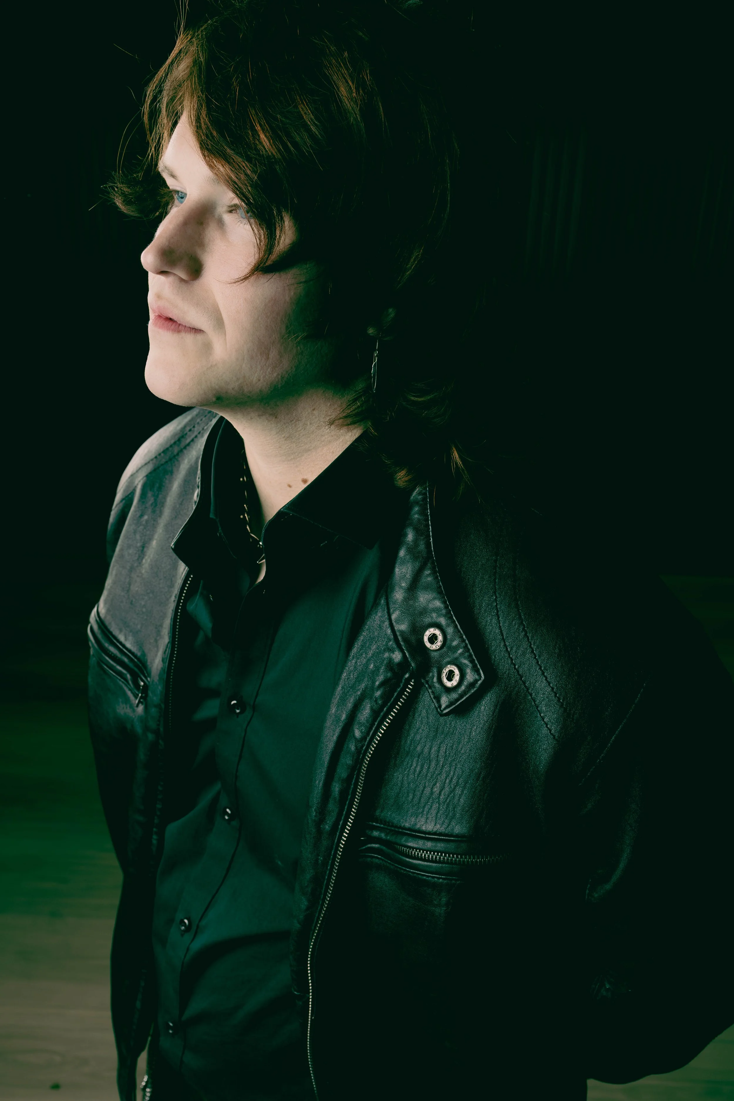 A young man with medium-length brown hair and fair skin, wearing a black leather jacket and black shirt, stands against a dark background, looking slightly left.