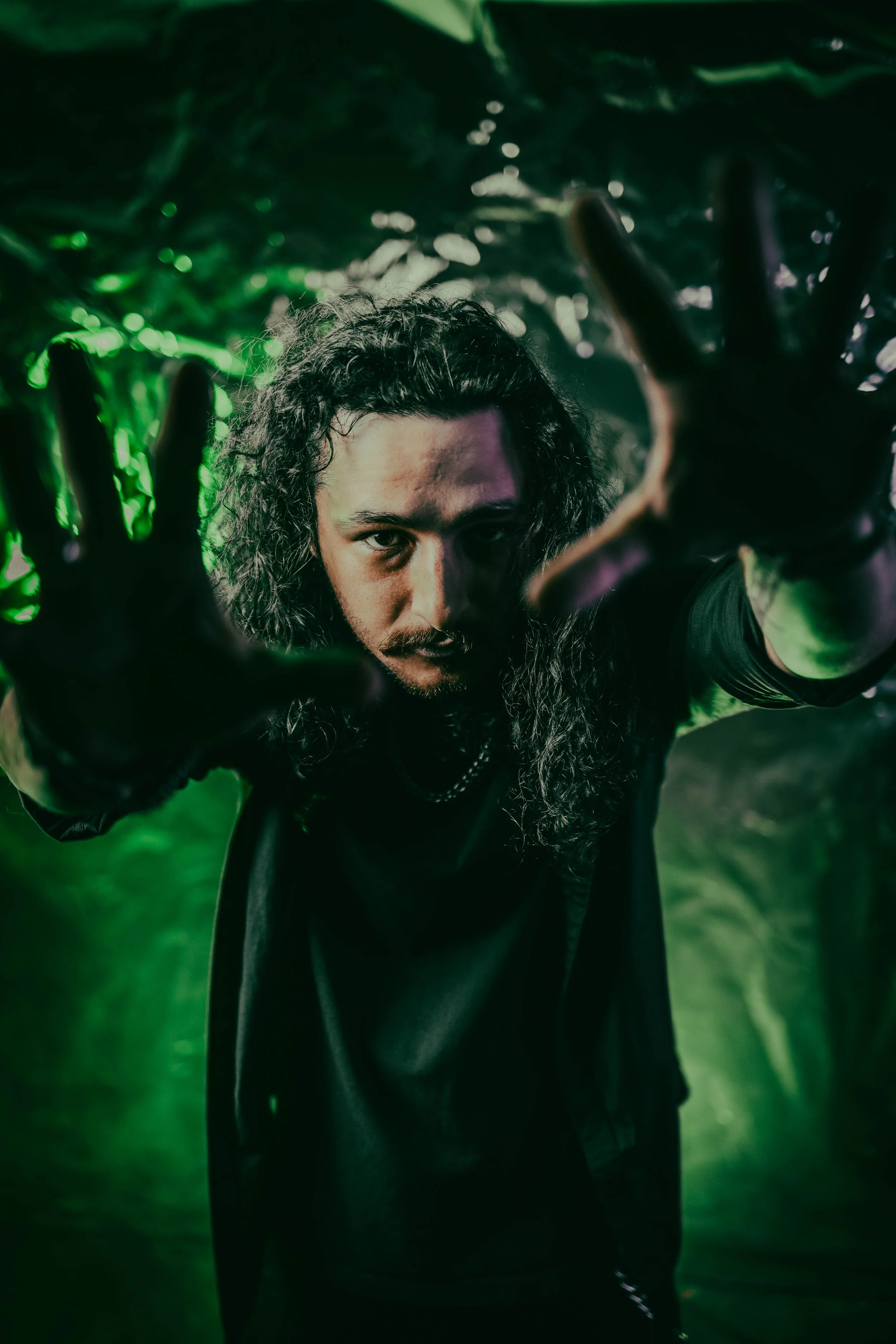 A man with long curly hair and a beard wearing black clothing reaches towards the camera with outstretched hands, in a dark green lit environment.