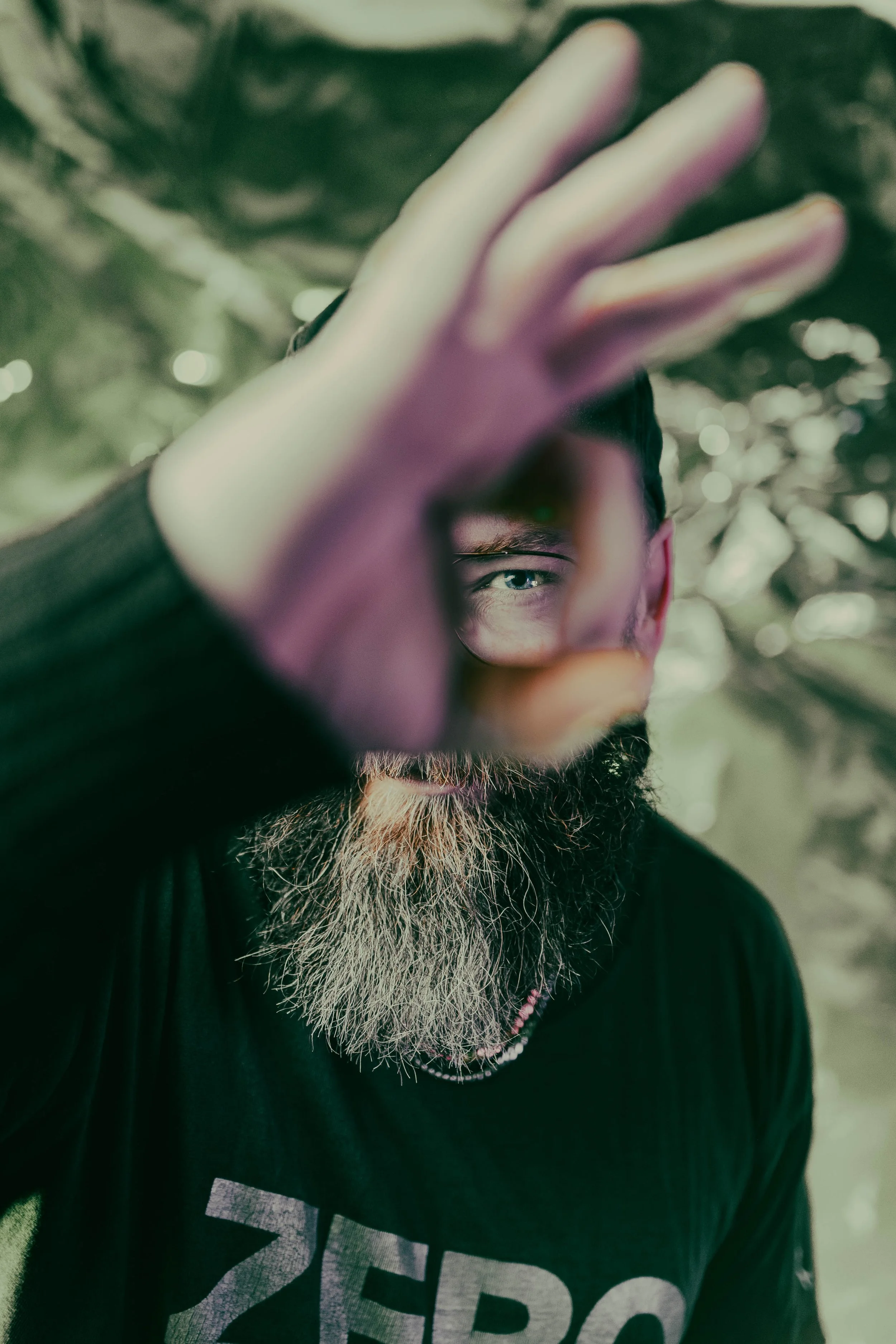 Person with a beard and glasses, covering part of their face with their hand, wearing a black shirt with gray text, outdoors with trees and dappled sunlight in the background.