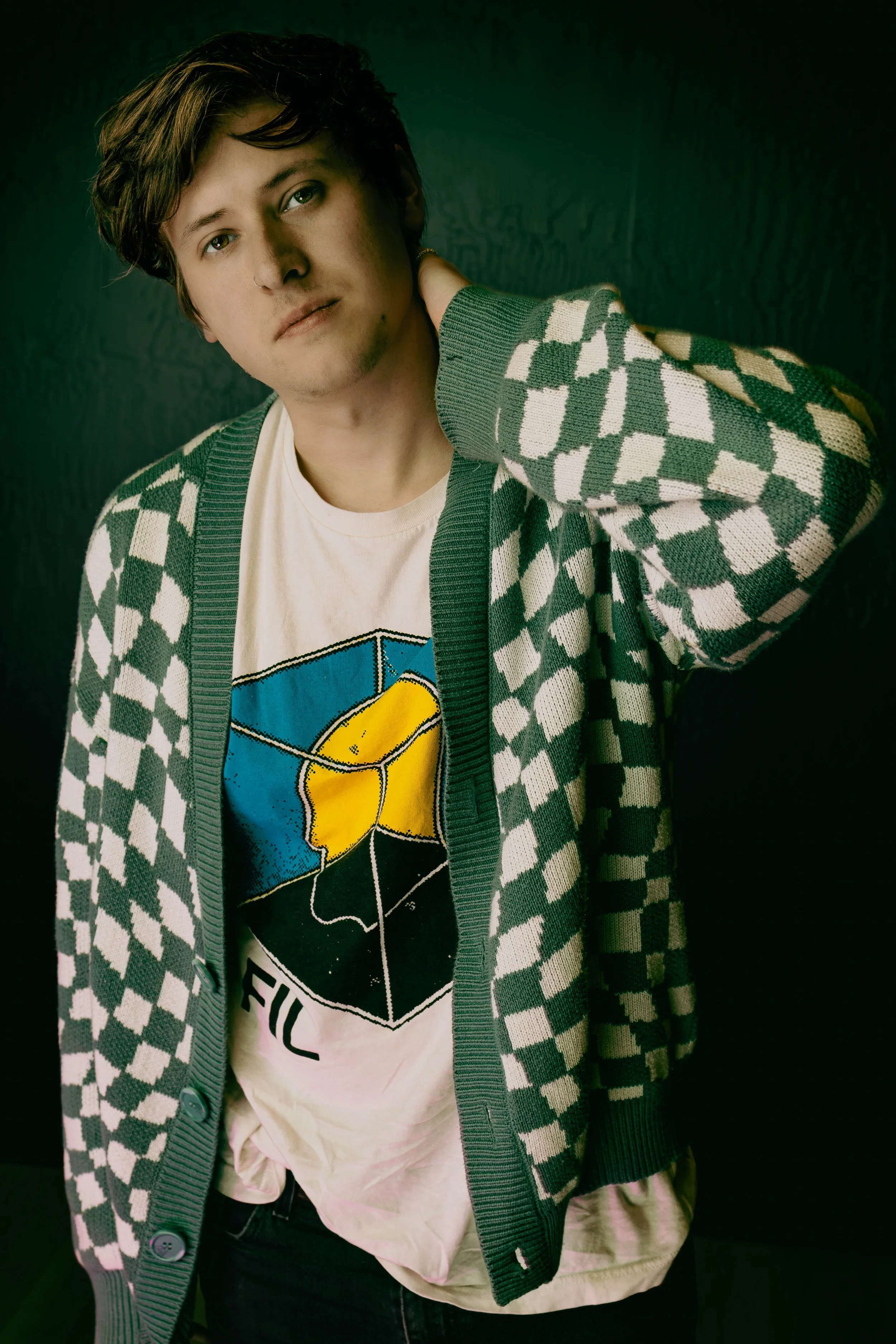 Young person with short brown hair, light skin, and a nose ring, wearing a green and white checkered cardigan over a graphic t-shirt featuring a Rubik's cube and the word 'FU' visible, standing against a dark background.