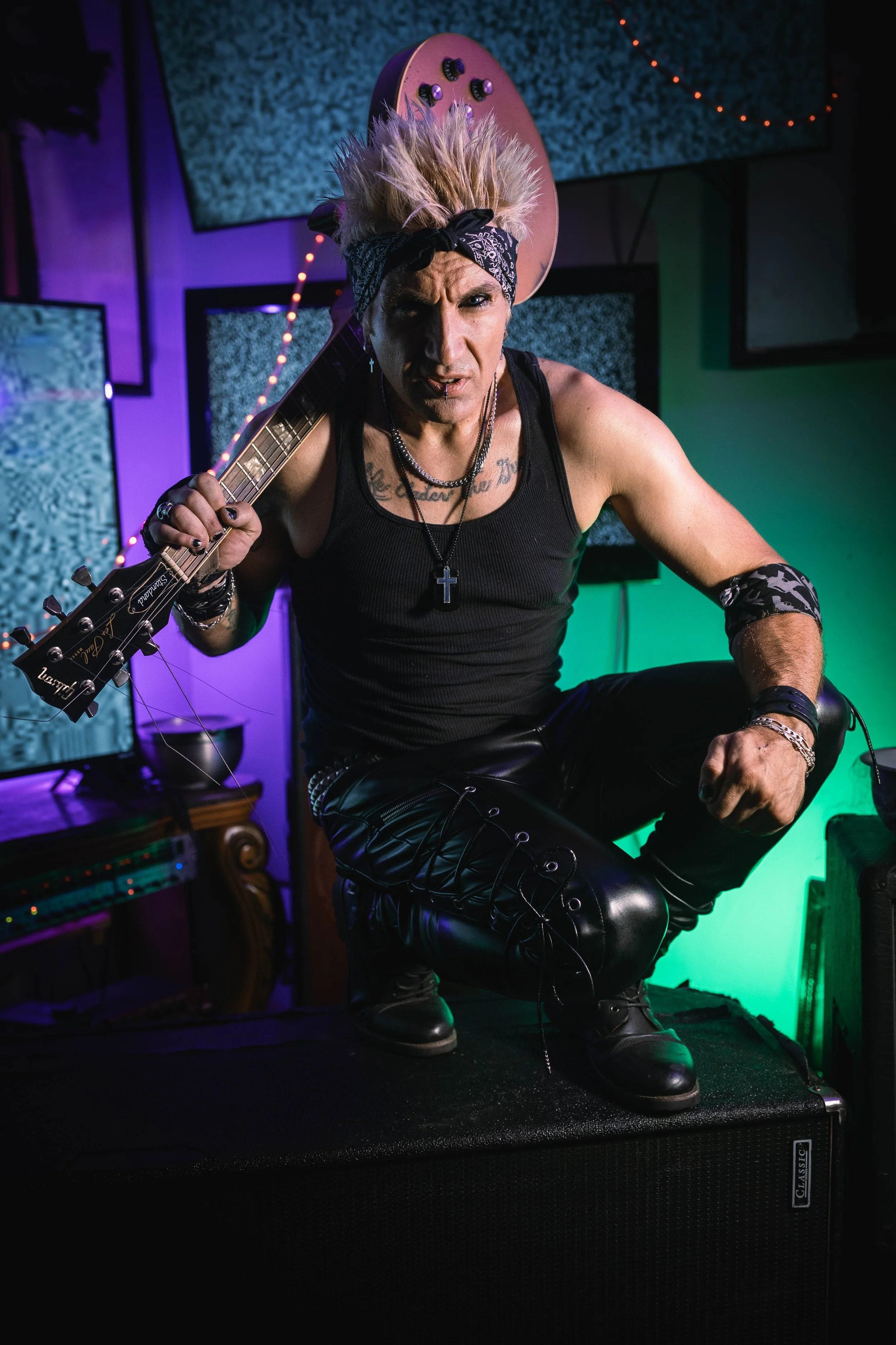 A musician with spiked blond hair, wearing a black bandana and black tank top, crouching atop a large speaker with a guitar strapped to his back, in a room with colorful lighting and TV screens.