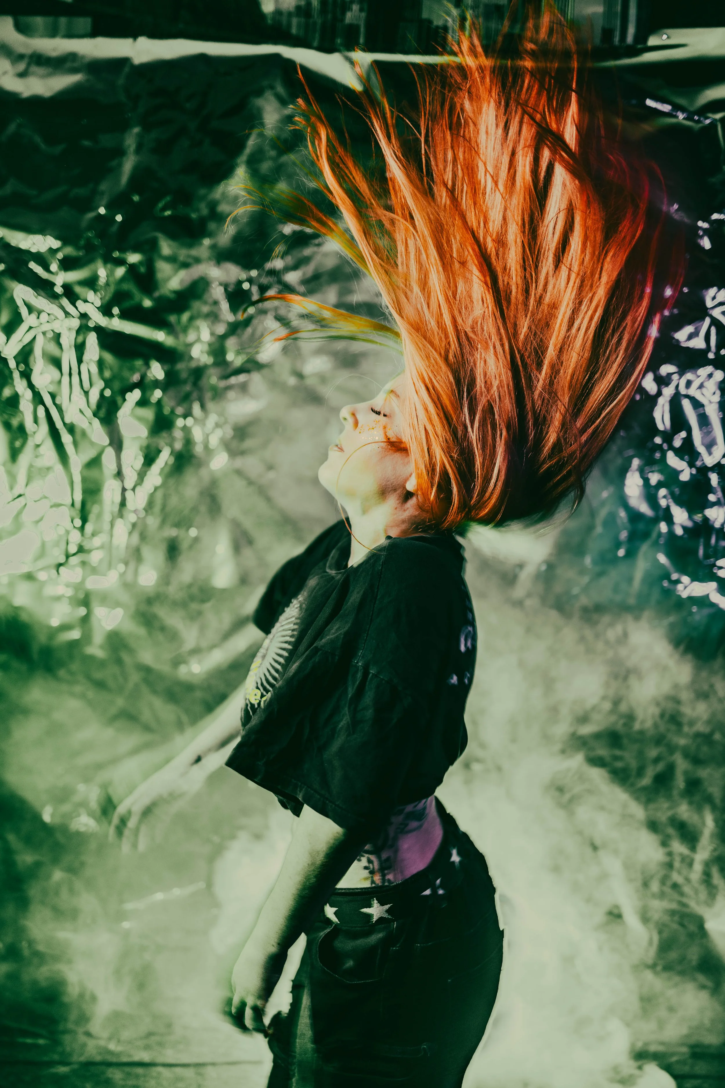 A young girl with bright red hair and a black shirt, standing outdoors near a white fence, with trees in the background. The image has a filter that gives it a greenish tint.
