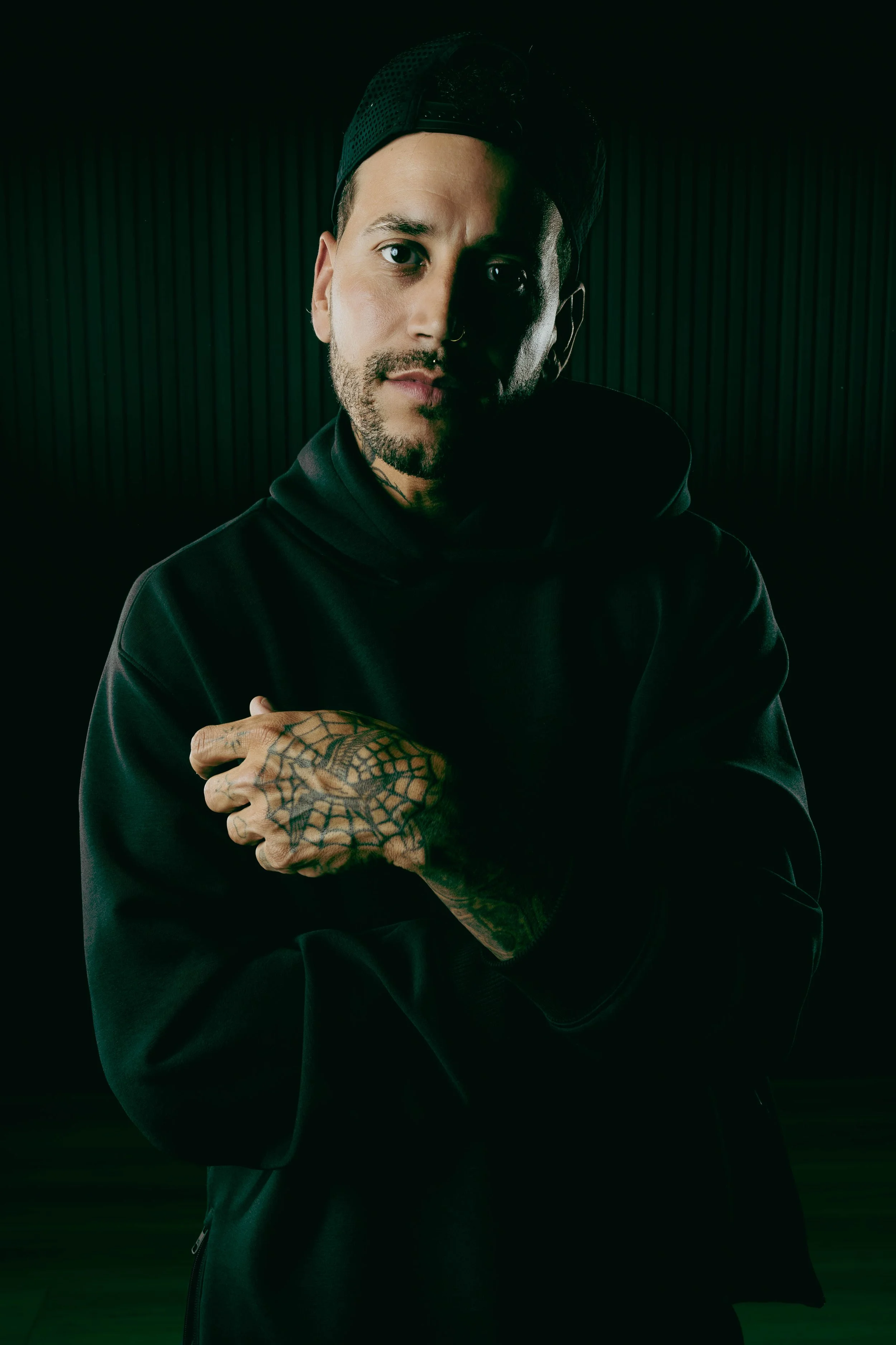 A man with a beard, tattoos on his hand, wearing a black hoodie and a black baseball cap backwards, posing in front of a dark, ribbed background.