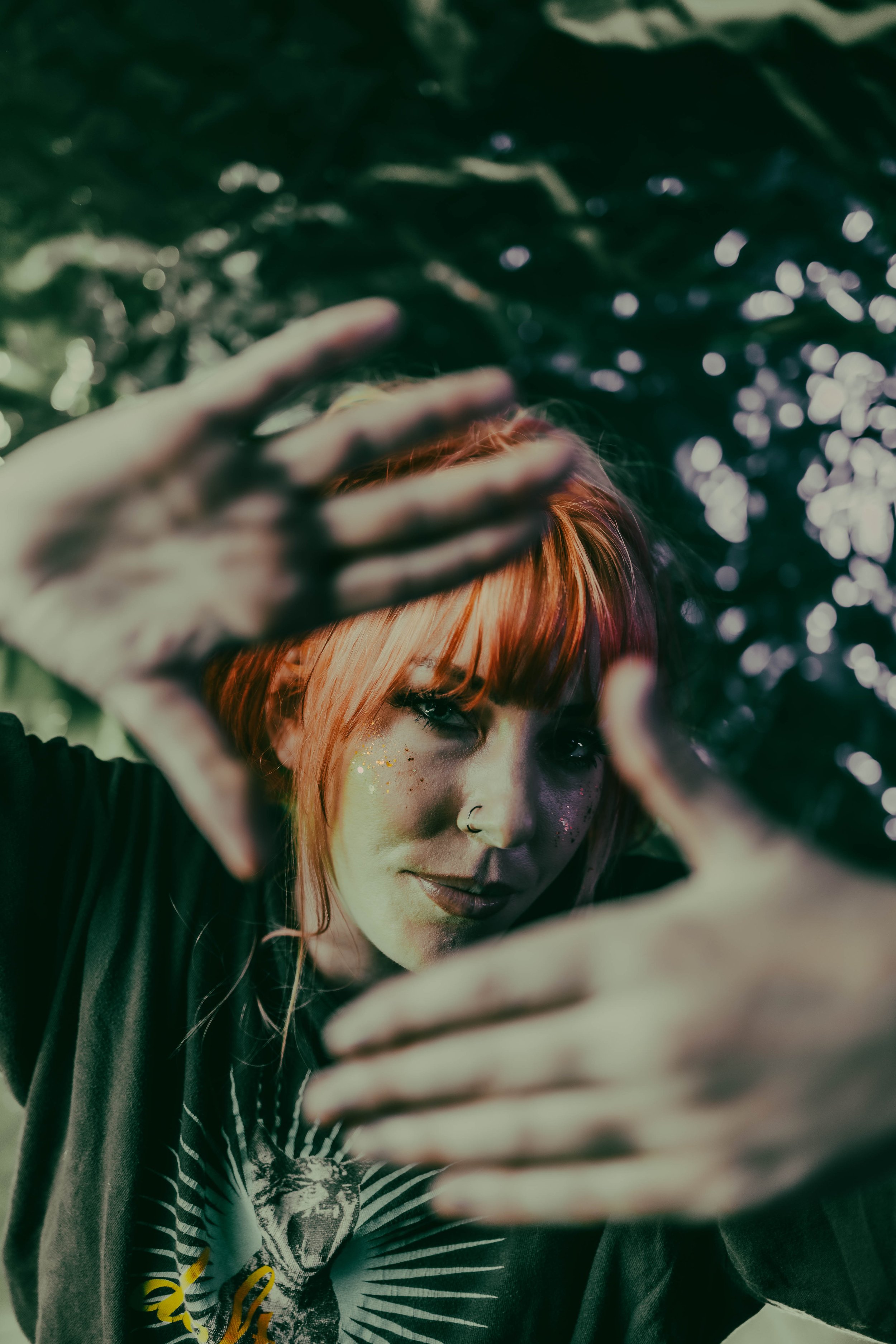 A person with orange hair and glitter on their face, wearing a black shirt with a graphic design, making a heart shape with their hand in front of their face.