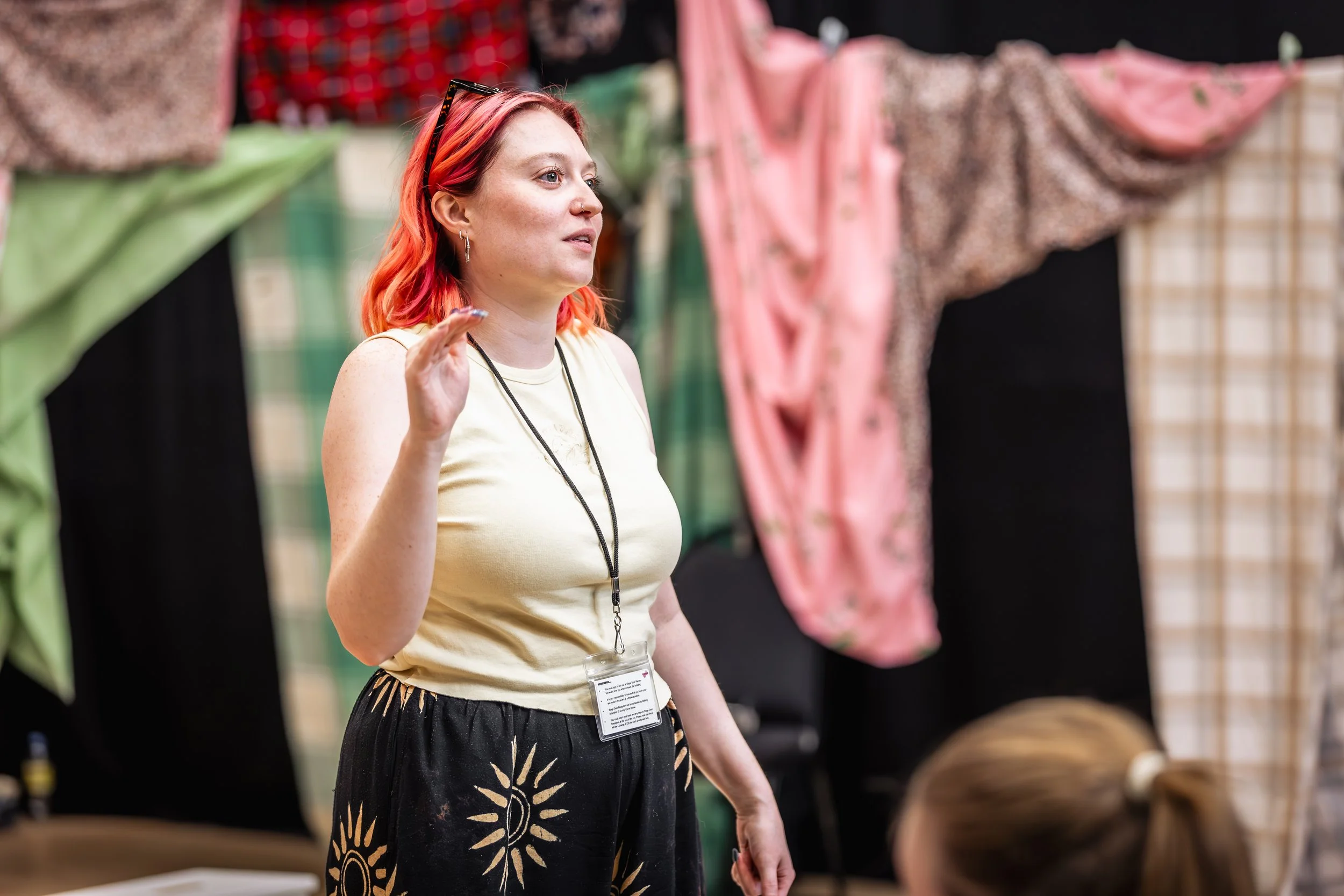 A woman with red hair, wearing a cream t-shirt, black trousers with sunflower pattern, glasses on her head, earrings, a nose ring, and a name badge, standing in front of hanging clothes which make up the set, giving notes in a rehearsal.