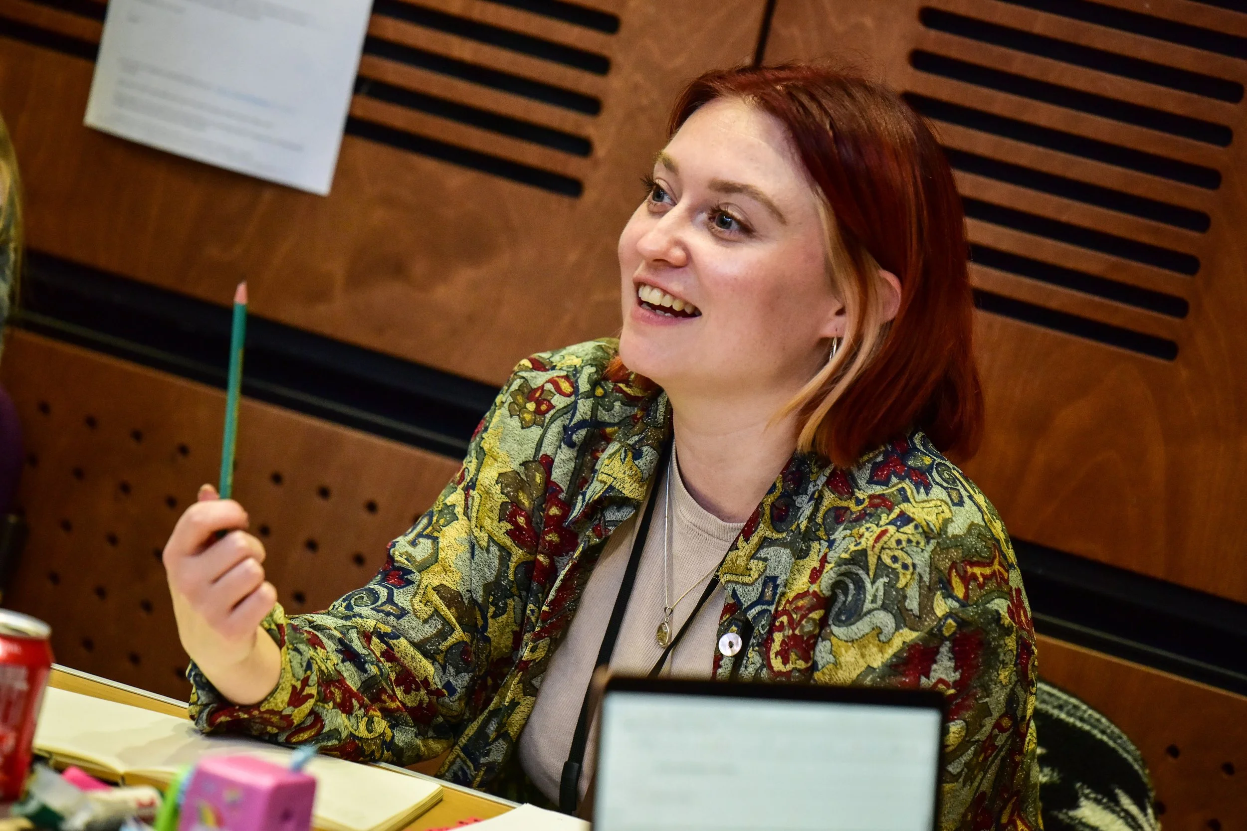 A woman with red hair smiling and holding a green pencil, sitting at a table with various colorful objects, in a room with wooden wall panels.