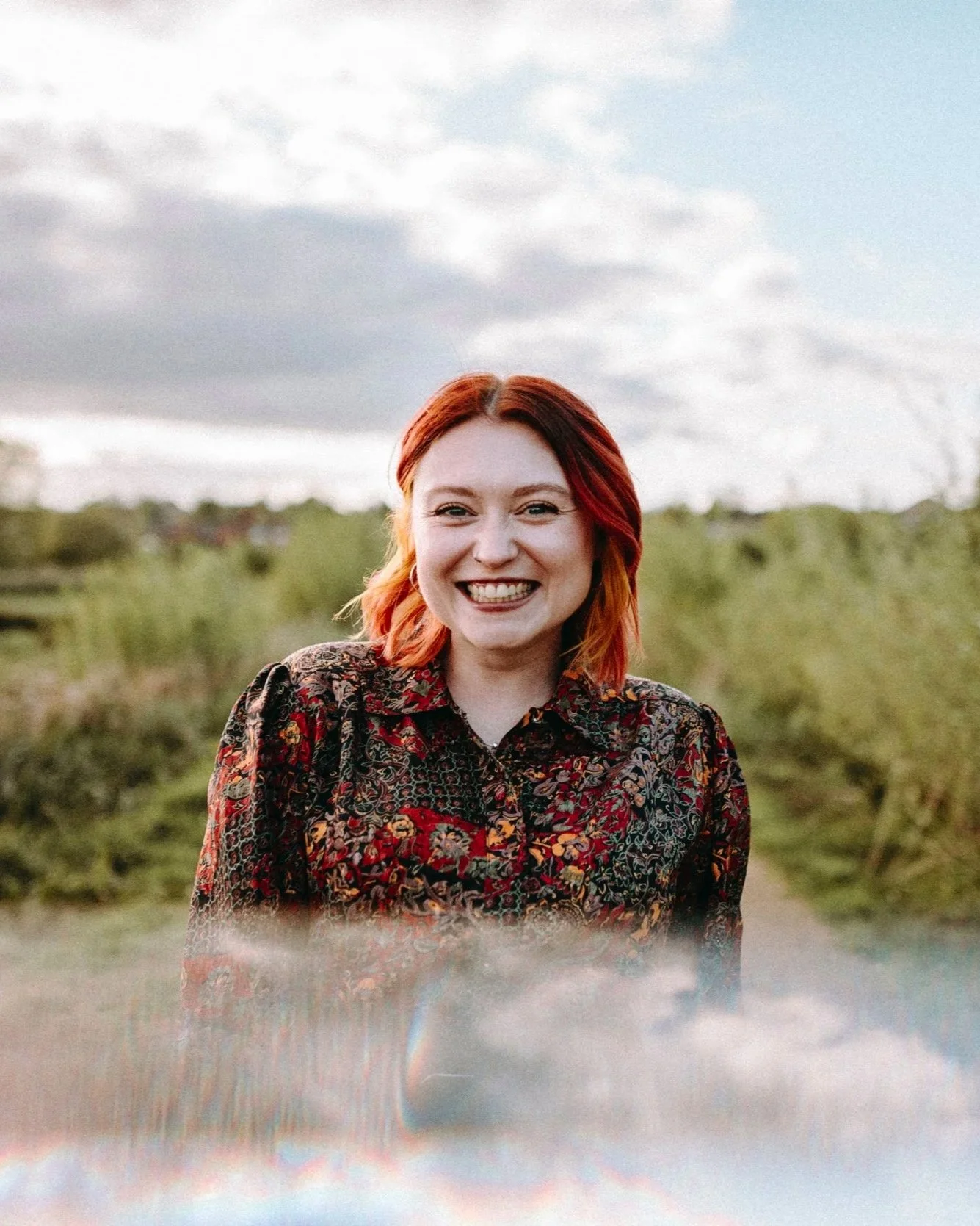 A woman with red hair smiling outdoors, standing on a grassy area with cloudy sky in the background.