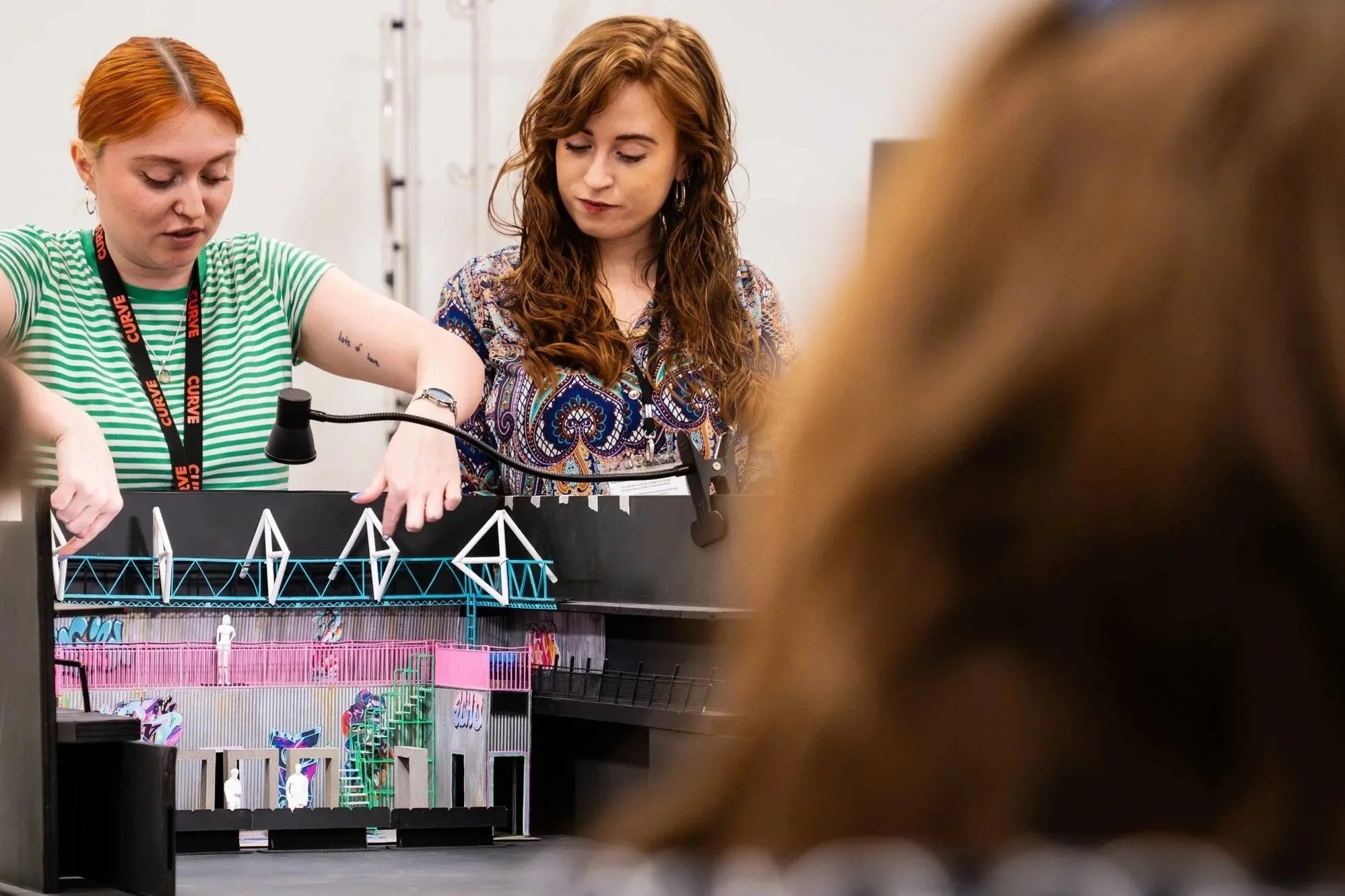 Two women working on a miniature model of a bridge with colorful structures, one adjusting the model while the other observes.