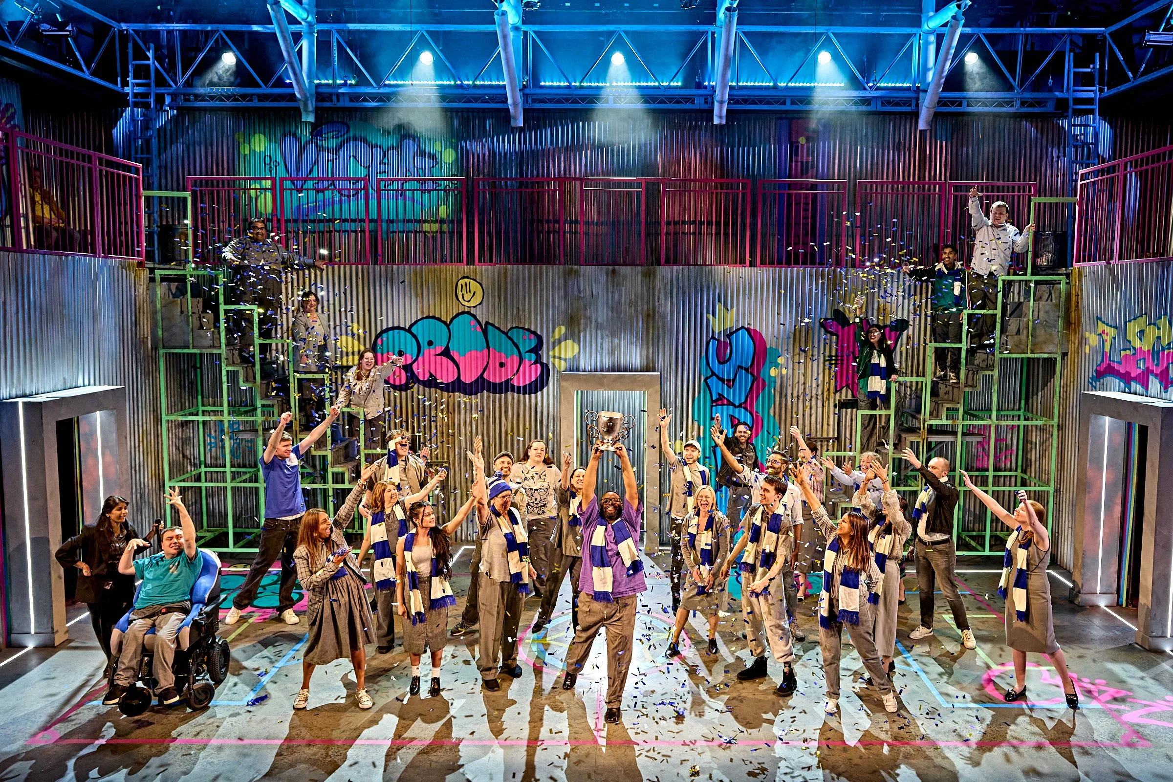 A large group of people celebrating on a stage with confetti, dramatic stage lighting and blue and white scarfs , in front of a colorful graffiti-style backdrop.