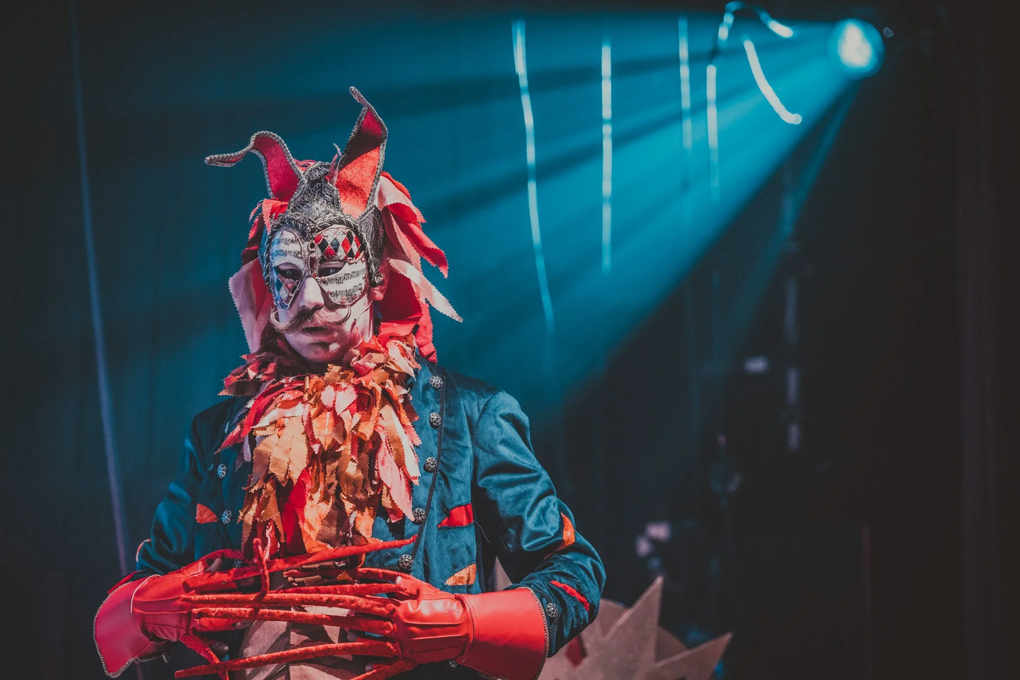 Person wearing a colourful beast costume with a mask, and horns, with his long beauty hands clasped together, in front of a dark background with dramatic blue stage lights.