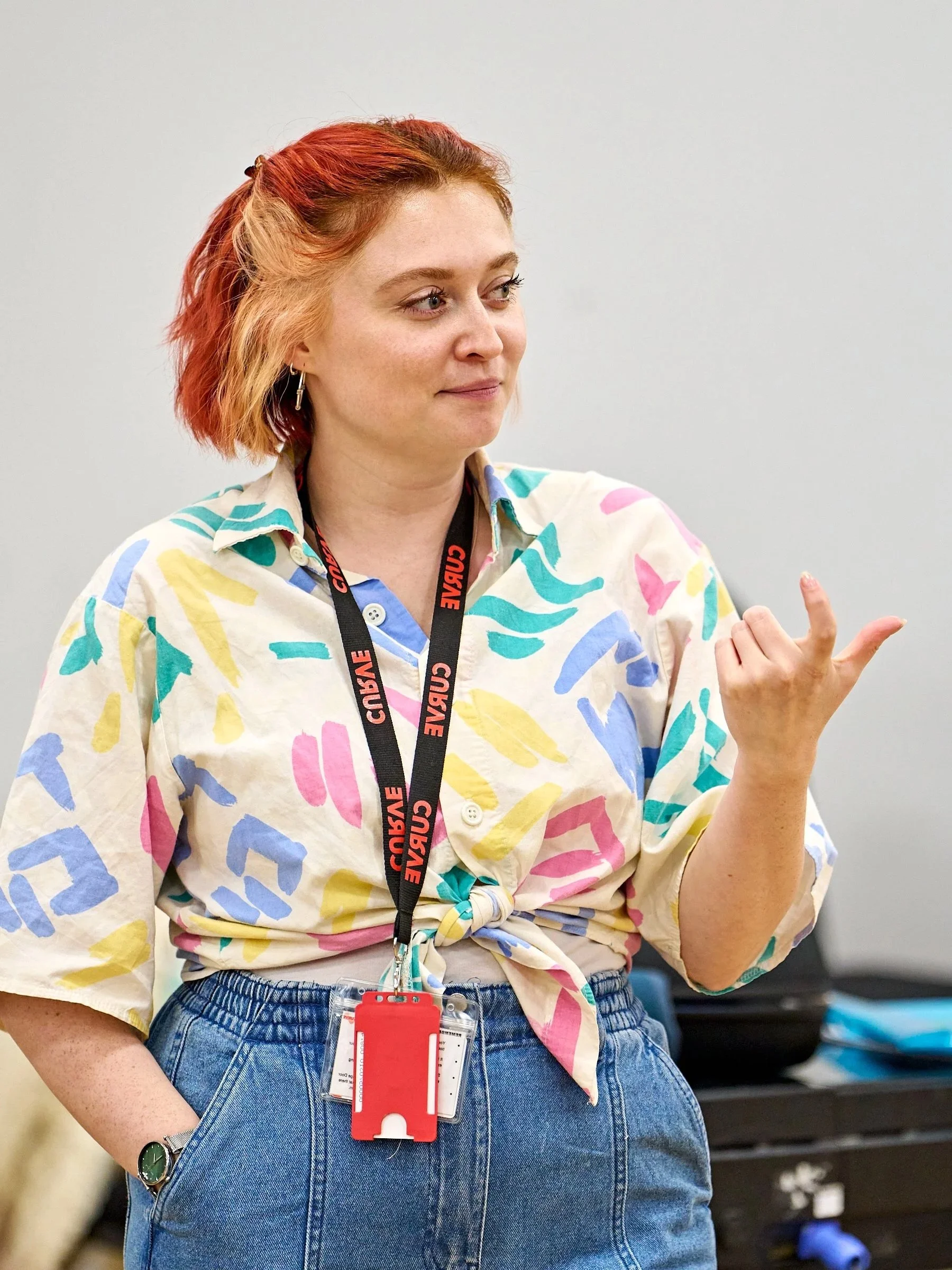 A woman with short red and blonde hair, wearing a colorful patterned shirt, blue jeans, and a wristwatch, standing indoors with a plain wall and some objects in the background, explains an idea to the room.