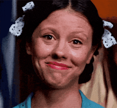 A woman with dark hair styled in braids and decorated with white lace hair accessories, smiling and wearing a blue top.