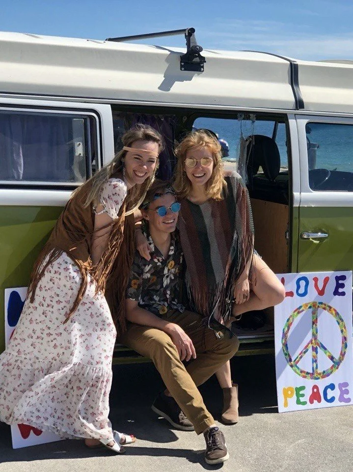 Three women smiling in front of a vintage camper van with a peace sign and love peace sign poster beside them, dressed in bohemian style outfits, by the waterfront.