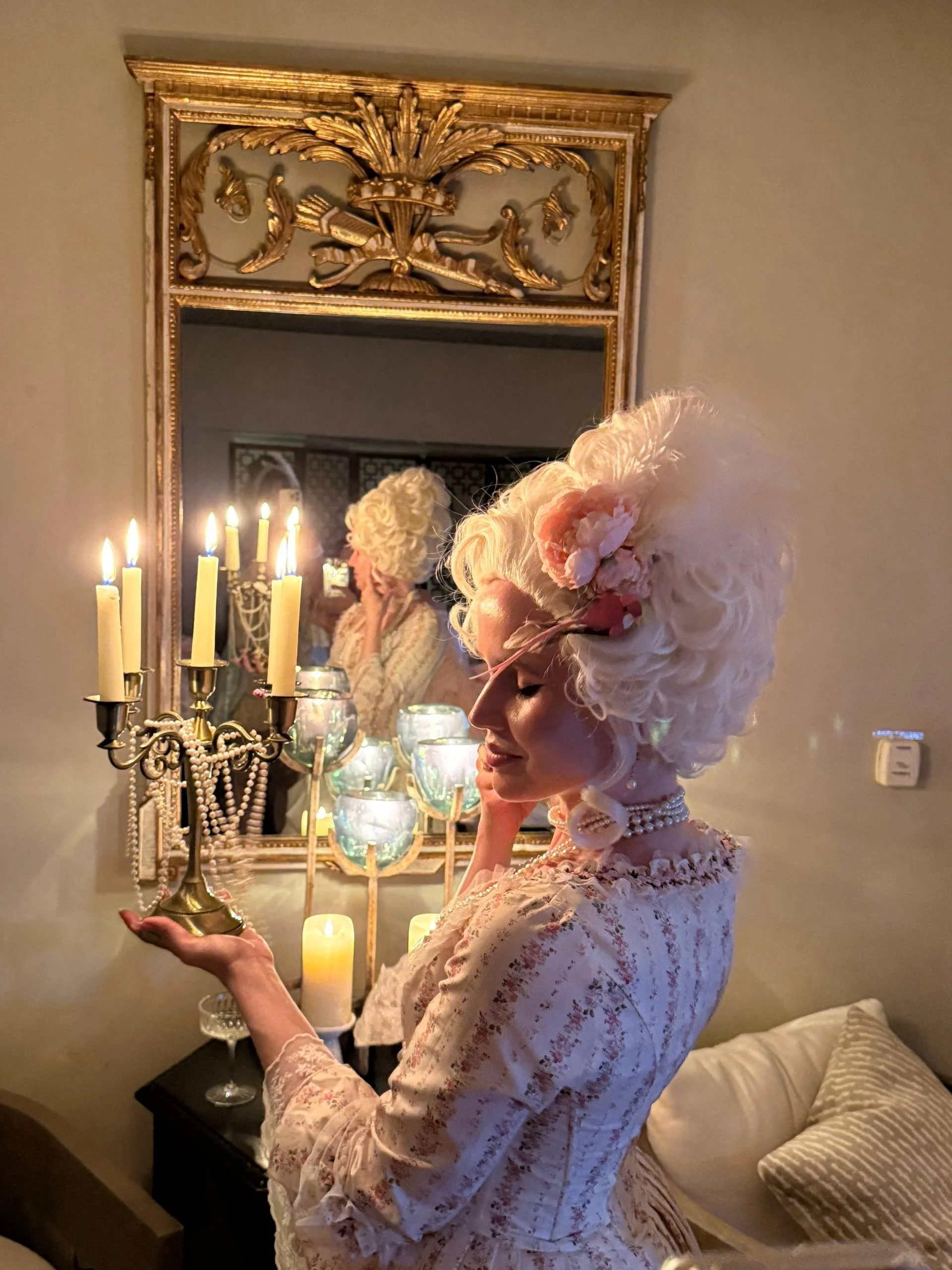 A woman dressed in vintage-style clothing with a floral pattern, wearing an elaborate feathered hat with large pink flowers, stands near a mirror, holding a candelabrum with lit candles, and a string of pearls around her neck. The scene is softly lit with warm candlelight.