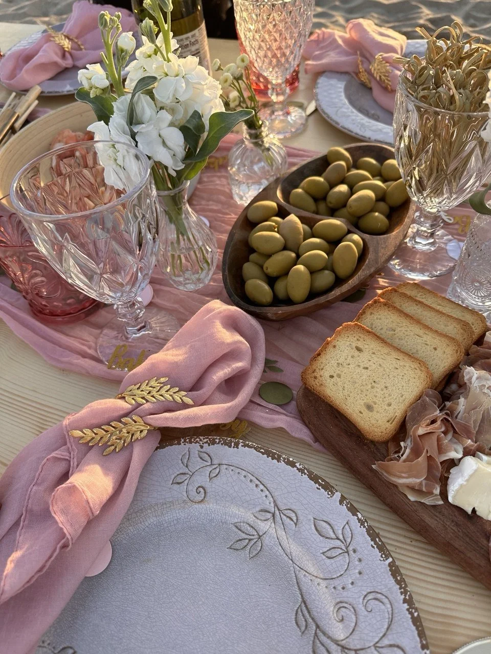 Elegant table setting with pink cloth napkins, crystal glasses, white flowers, green olives in a wooden bowl, slices of bread, and prosciutto and cheese on a serving board.