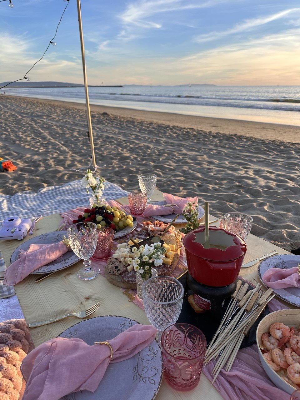 Beach picnic setup with a table of food and drinks, pink napkins, and glasses, overlooking the ocean during sunset.