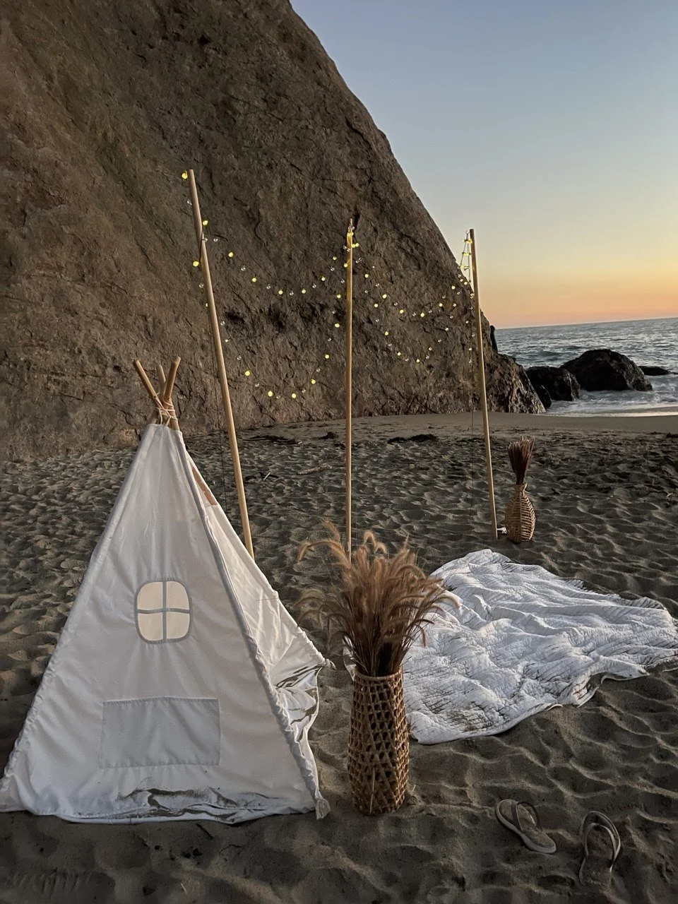 Beach setup with a small white tent, wicker vases containing pampas grass, string lights hanging from wooden poles, and a blanket on sand near rocks and ocean at sunset.