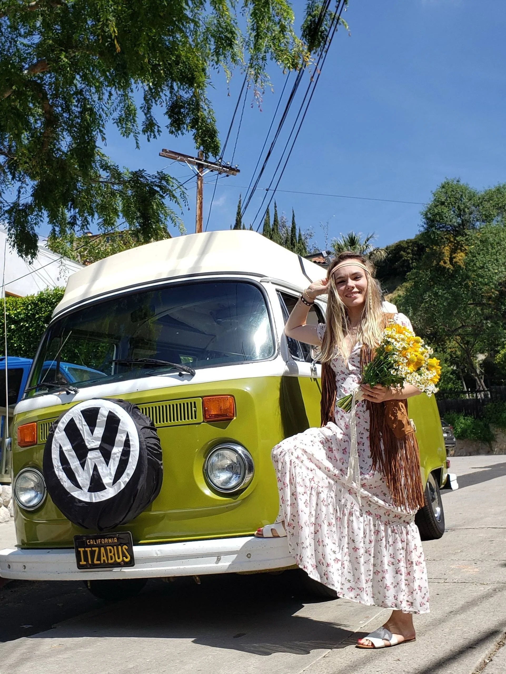 A young woman with long blonde hair wearing a white floral dress and a brown fringe shawl holding a bouquet of yellow and white flowers, standing next to a vintage green Volkswagen van with a California license plate reading "TIZZBUS" and a tire cover with the Volkswagen logo, outdoors on a sunny day with trees, blue sky, and power lines in the background.
