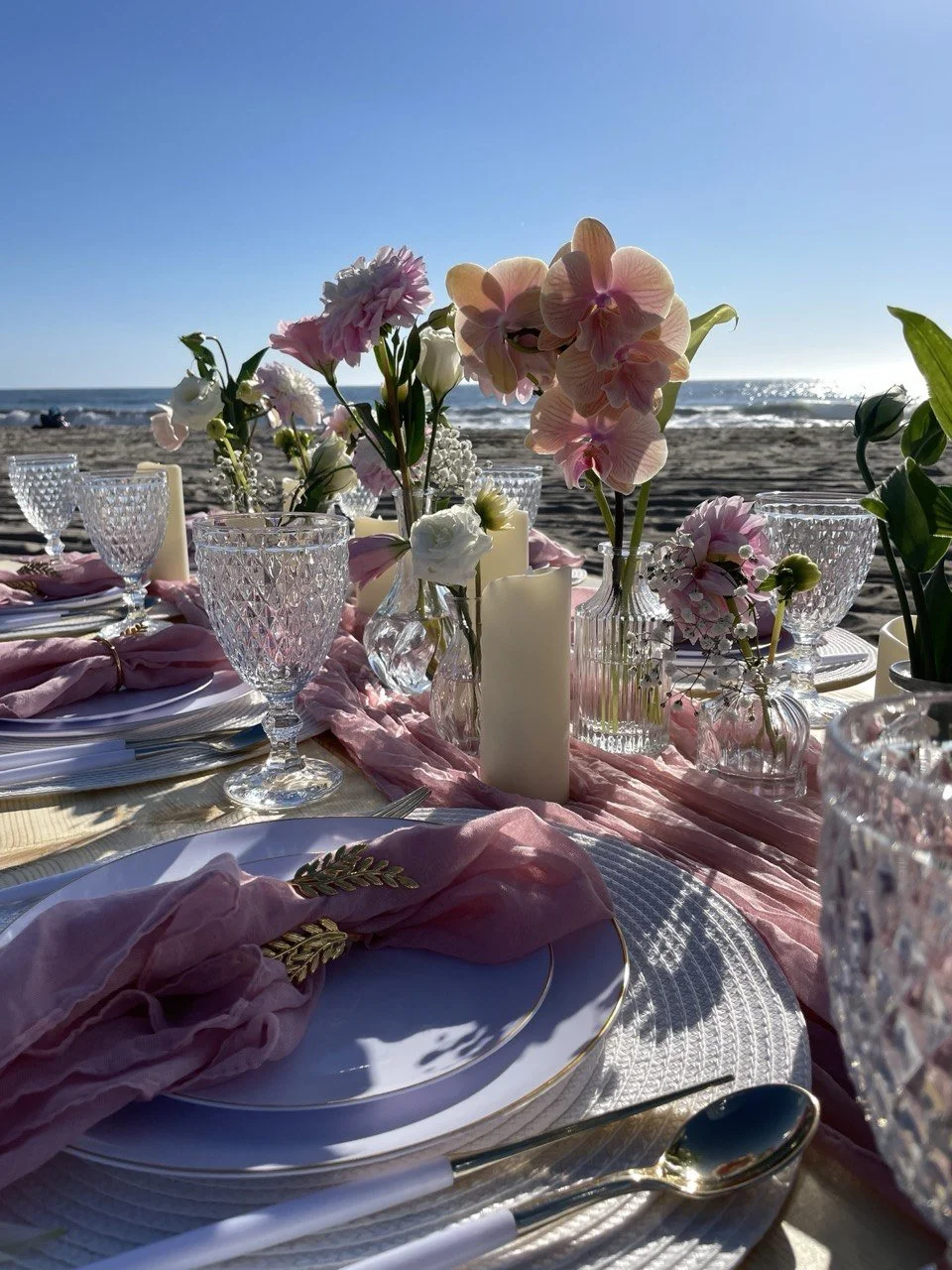 Beachside table setup with pink and white flowers, candles, crystal glassware, and fine dinnerware, overlooking the ocean at sunset.