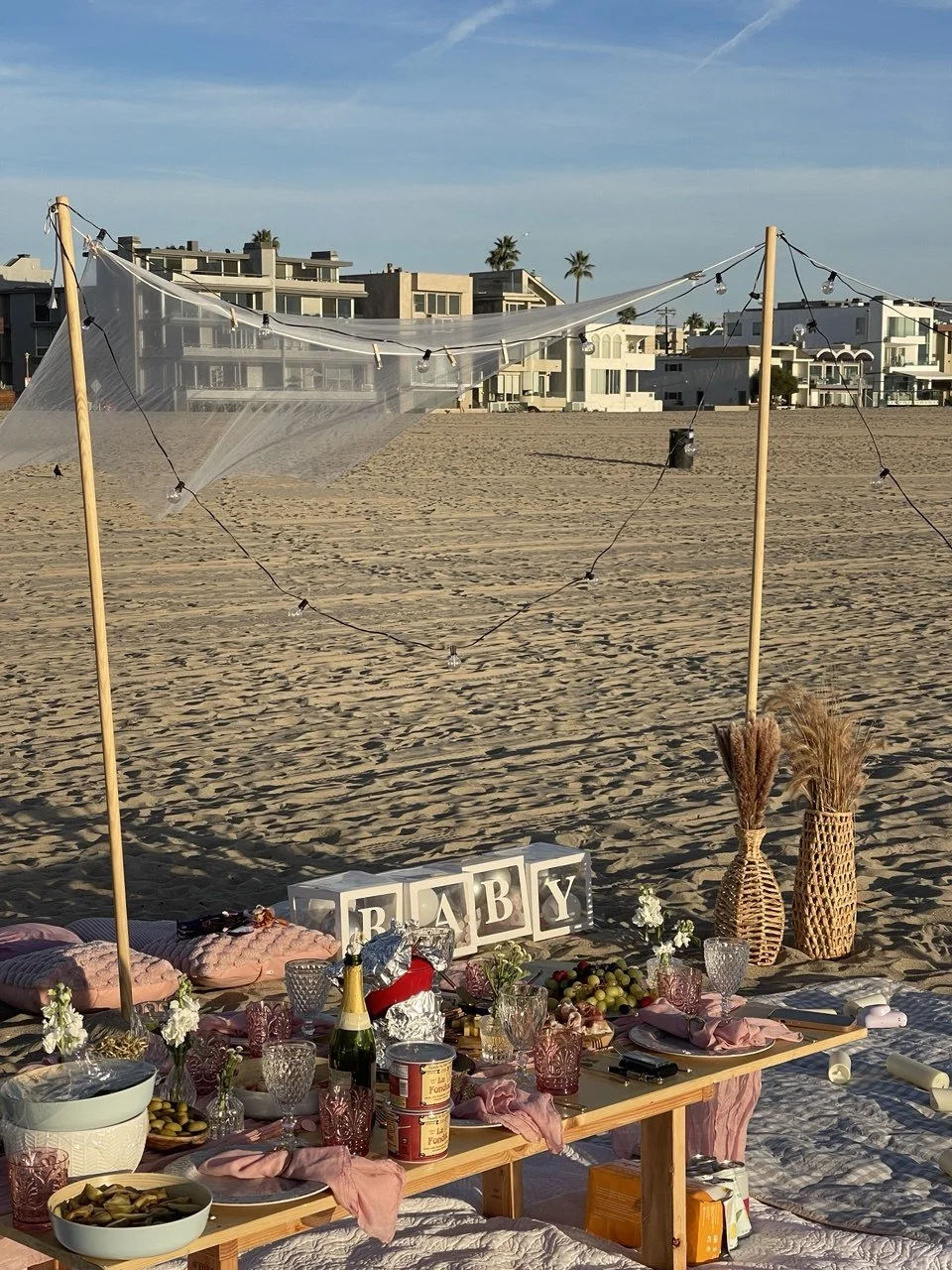 Beach picnic setup with a wooden table filled with food, drinks, and tableware, decorated with pink napkins and vases, under a string light and sheer fabric canopy, with a sign spelling 'BABY' in block letters, against a background of residential buildings and palm trees on a sandy beach.