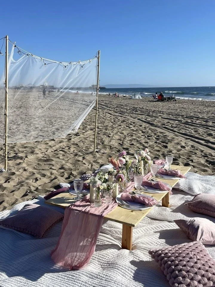 Beachside picnic setup with low tables, pink cloth, candles, and flowers on a white blanket, with ocean, sand, and a clear blue sky in the background