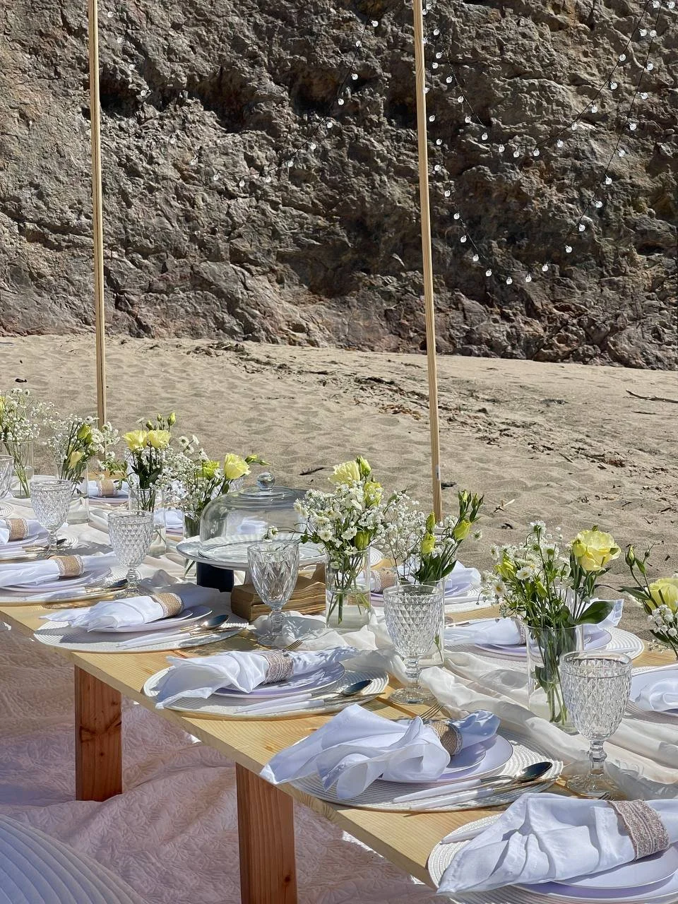 A beachside dining setup with a long wooden table decorated with white napkins, glassware, and centerpieces of white and yellow flowers in glass vases, set against a sandy beach and rocky cliff background.