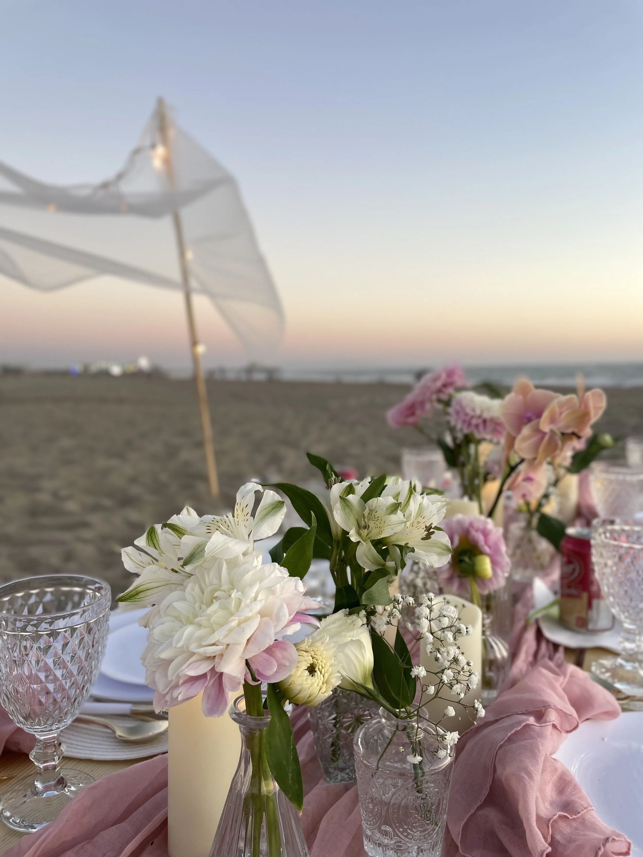 A close-up view of a floral centerpiece on a table at an outdoor beachside setting, with a blurred sunset sky in the background and a white patio umbrella.