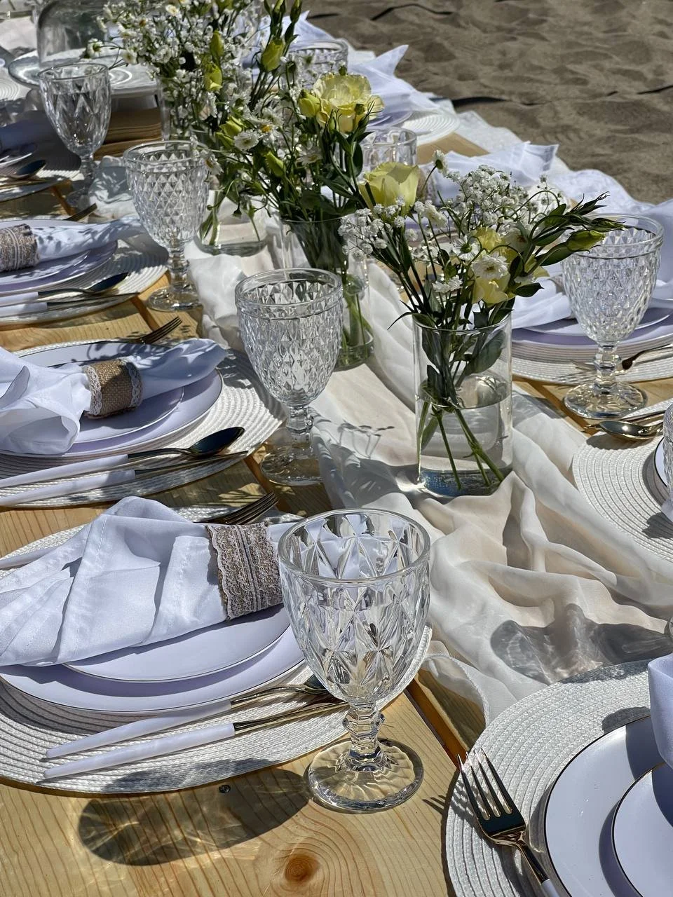 Table set for a formal outdoor event with white plates, silverware, crystal glasses, white napkins, and a floral centerpiece of white and pale yellow flowers, on a white table runner on a wooden table with a sandy background.