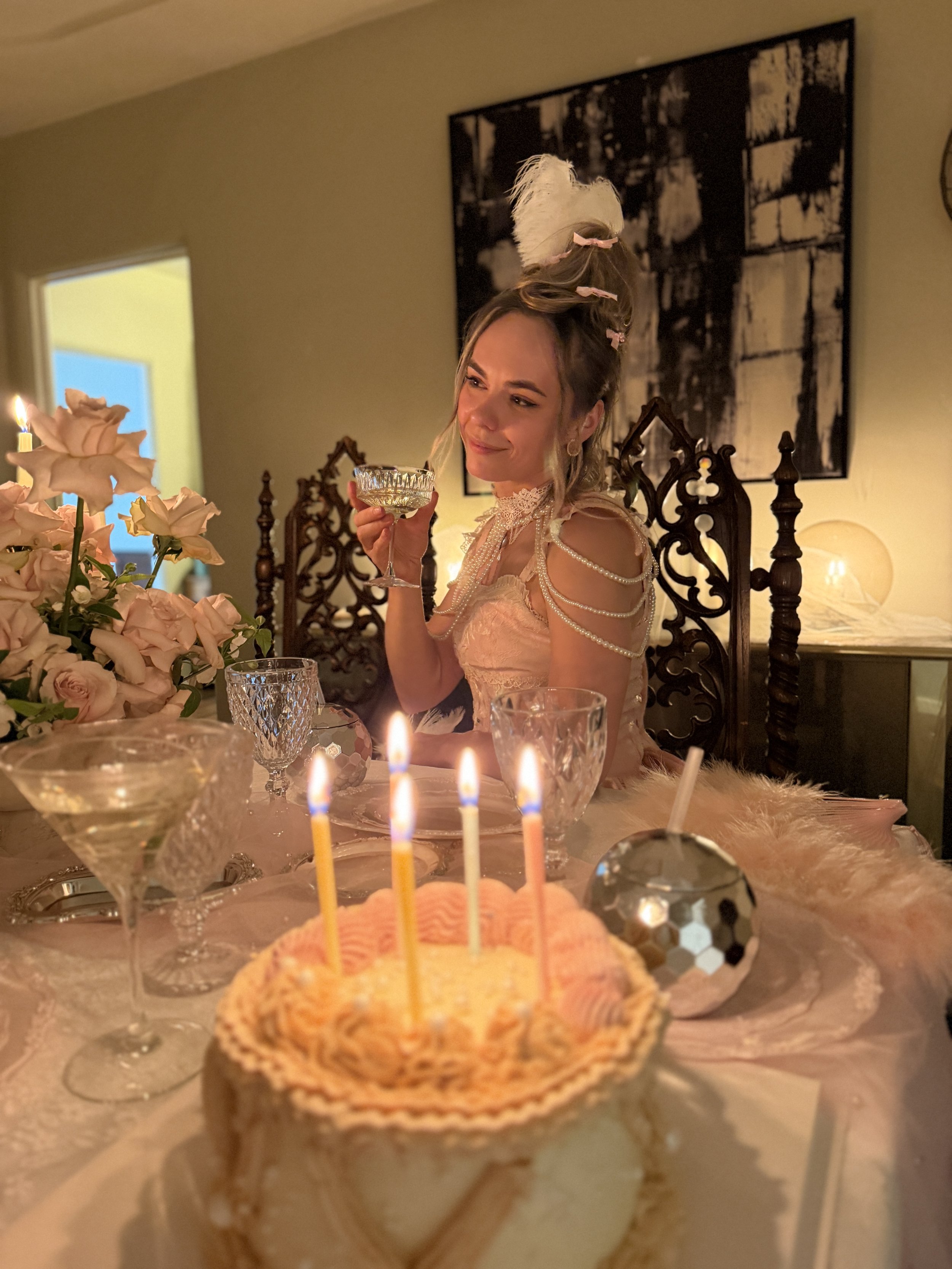 A woman dressed in vintage or Victorian-inspired attire holding a glass at a birthday celebration with candles on a cake, floral arrangements, and candles on the table.