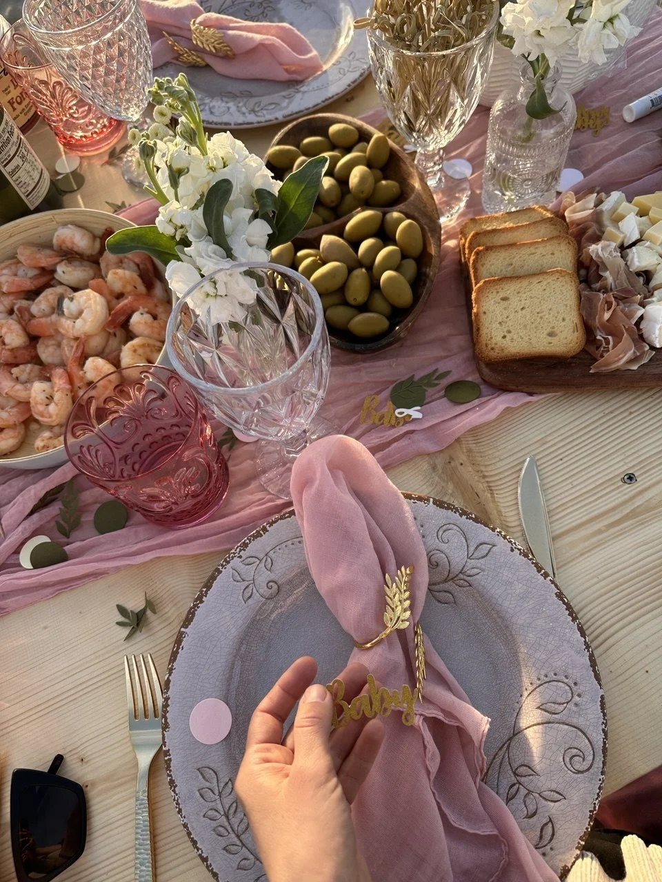 A decorated table set for a celebration with pink and gold accents, including a pink napkin with a "Baby" name tag, flowers, shrimp, olives, sliced bread, cheese, and various glassware.