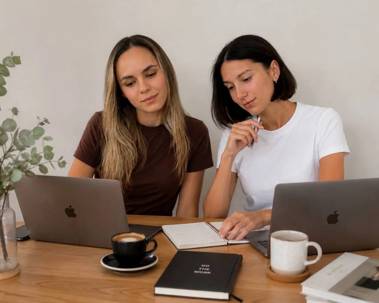 Two women working together at a wooden table with laptops, notebooks, coffee, and a plant in a bright room.