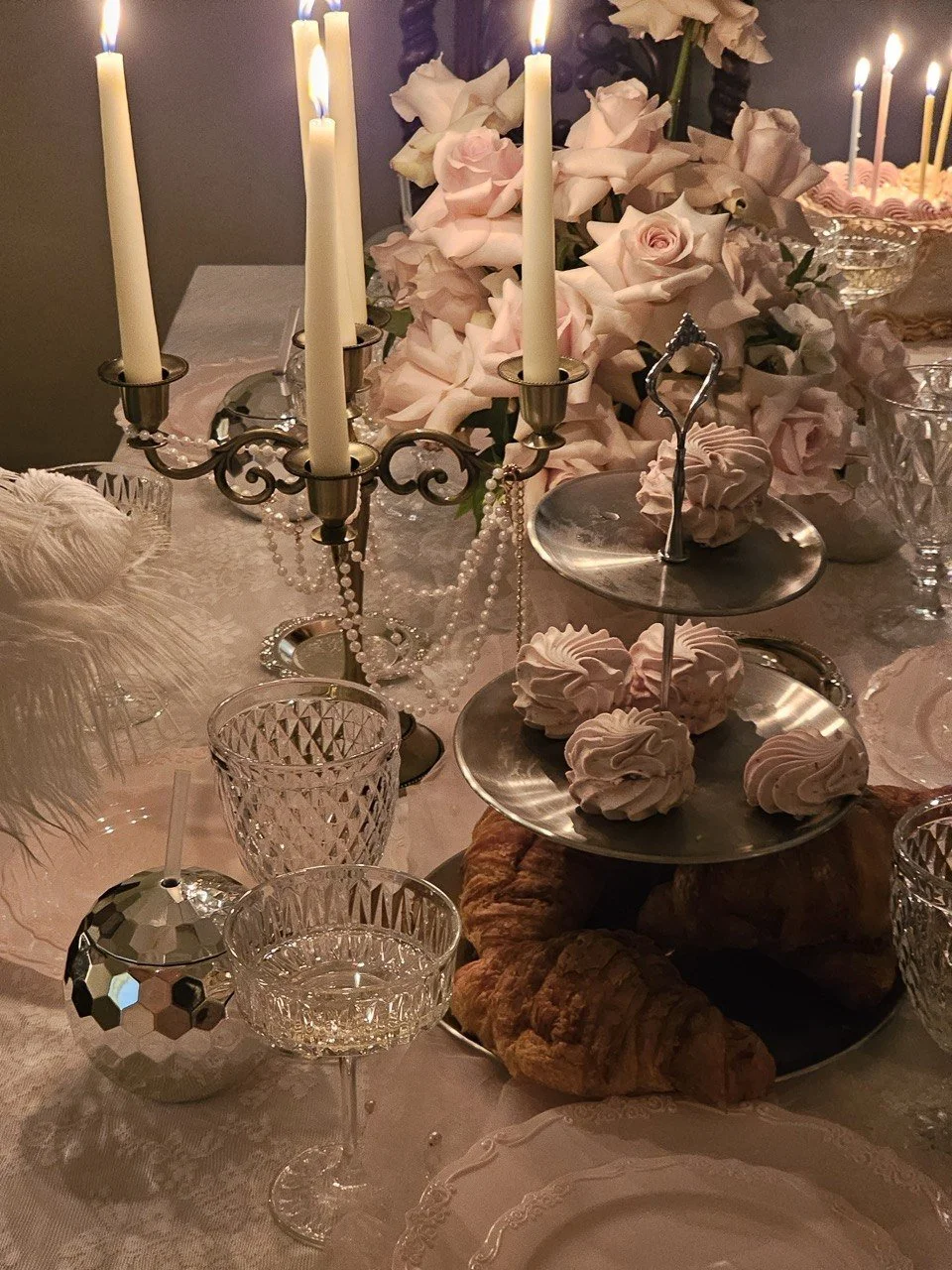 Elegant table setting with lit white candles in candleholders, a large floral arrangement of pink and white roses, a tiered tray with pink meringue cookies, croissants, glassware, and decorative elements, all on a lace tablecloth.