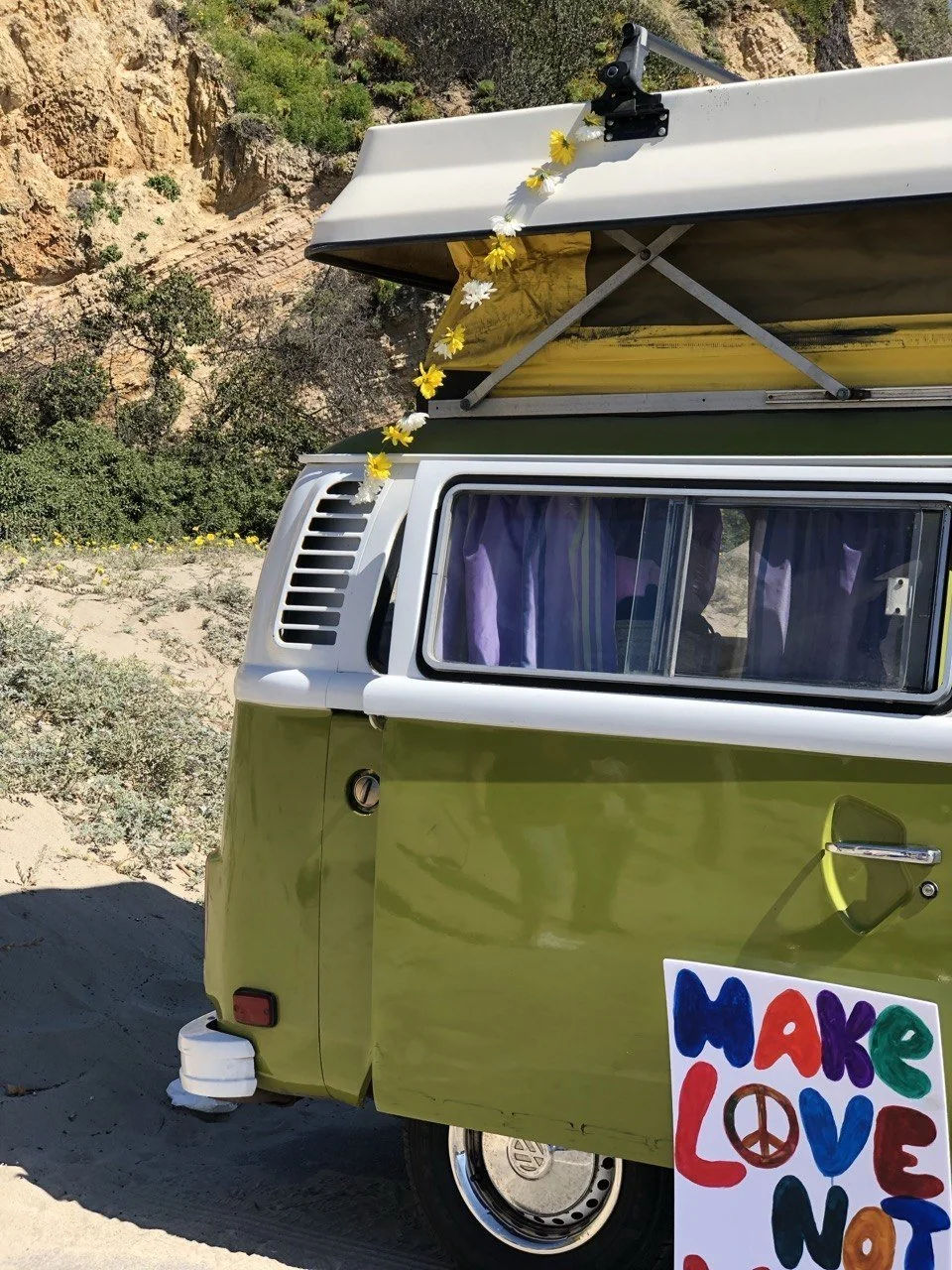 A vintage green and white Volkswagen camper van with a yellow roof, parked on a sandy area with bushes and rocks in the background. There is a sign in front of the van with a colorful message that says, 'Make Love Not War,' adorned with a peace symbol.