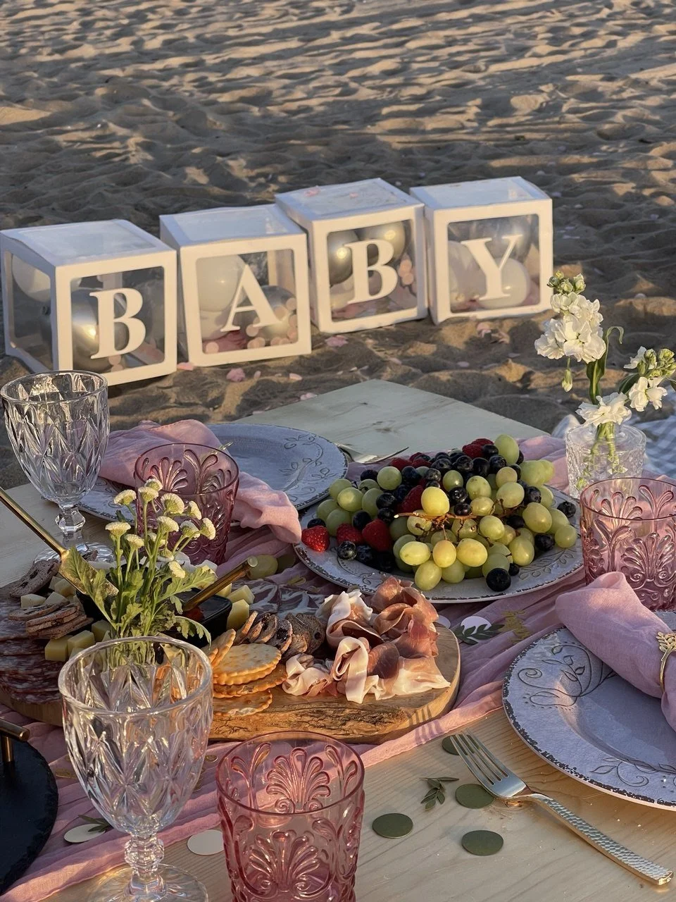 Beachside baby shower with a table set with plates, glasses, flowers, and a cheese platter, with illuminated baby blocks spelling 'BABY' in the background.