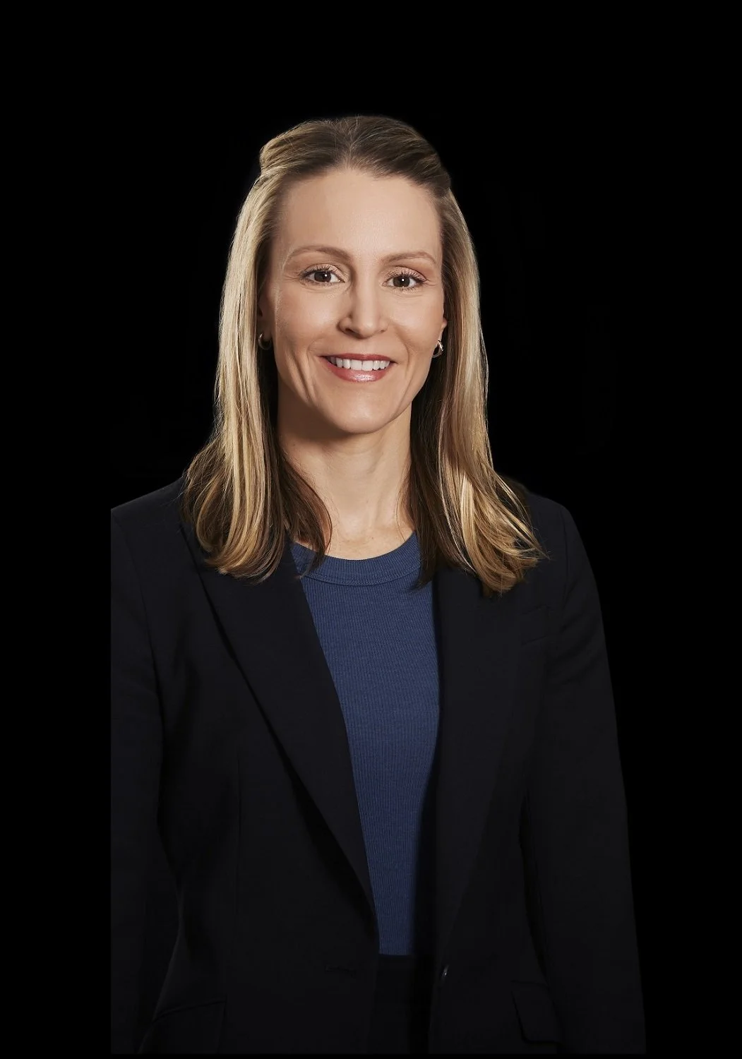 Professional woman with shoulder-length blonde hair, wearing a navy blazer and blue top, smiling against a black background.