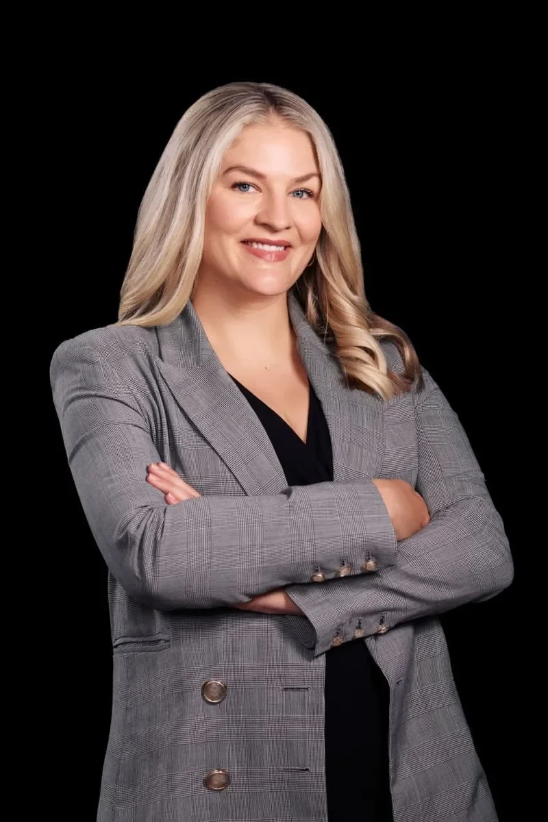 A woman with blonde hair smiling, arms crossed, wearing a gray plaid blazer over a black top, standing against a black background.