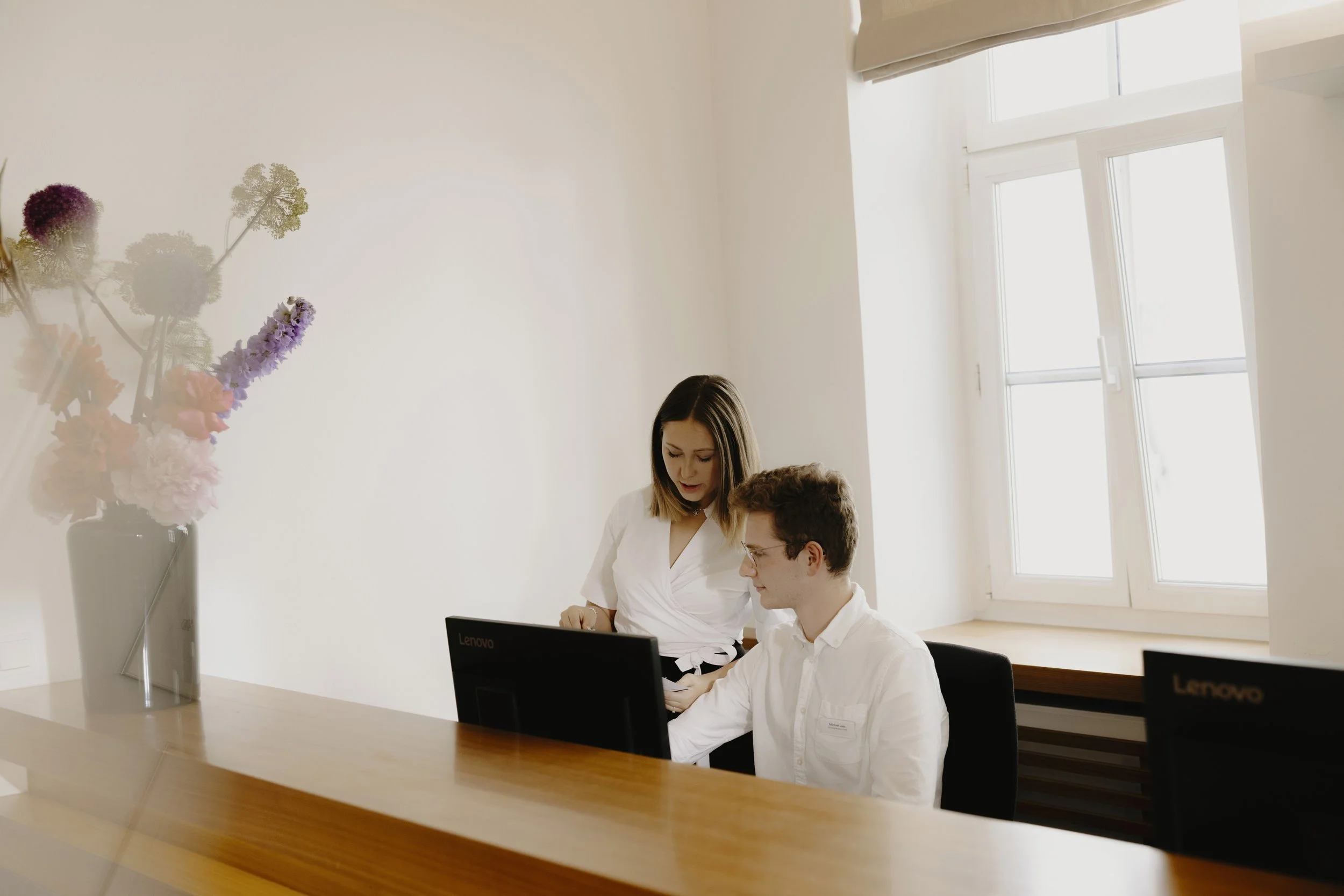 Drei Menschen sitzen an einem Tisch mit Laptops in einem hellen Raum. Vorne eine Vase mit bunten Blumen, im Hintergrund ein Fenster, das Tageslicht ins Zimmer lässt.