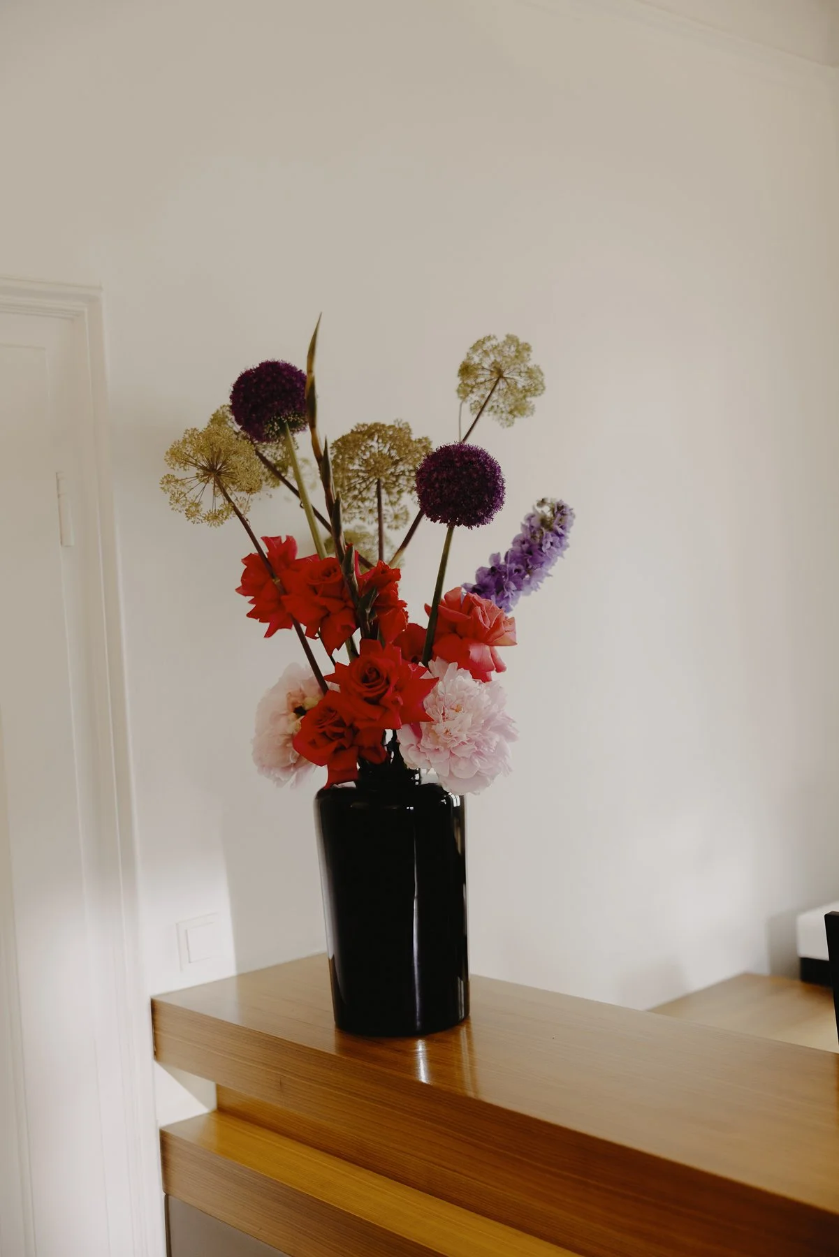 Blumenstrauß in schwarzer Vase auf einem Holztisch, vor einer weißen Wand.