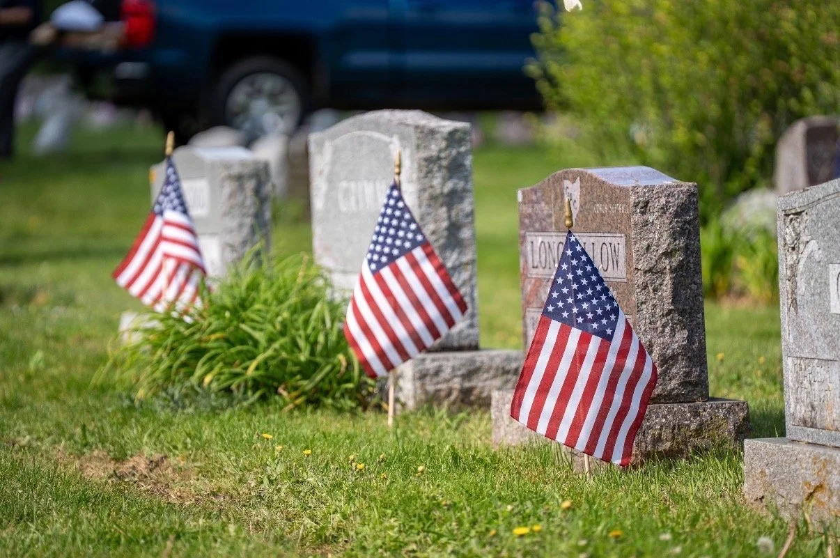 Four graves marked with headstones and decorated with small American flags in a grassy cemetery.