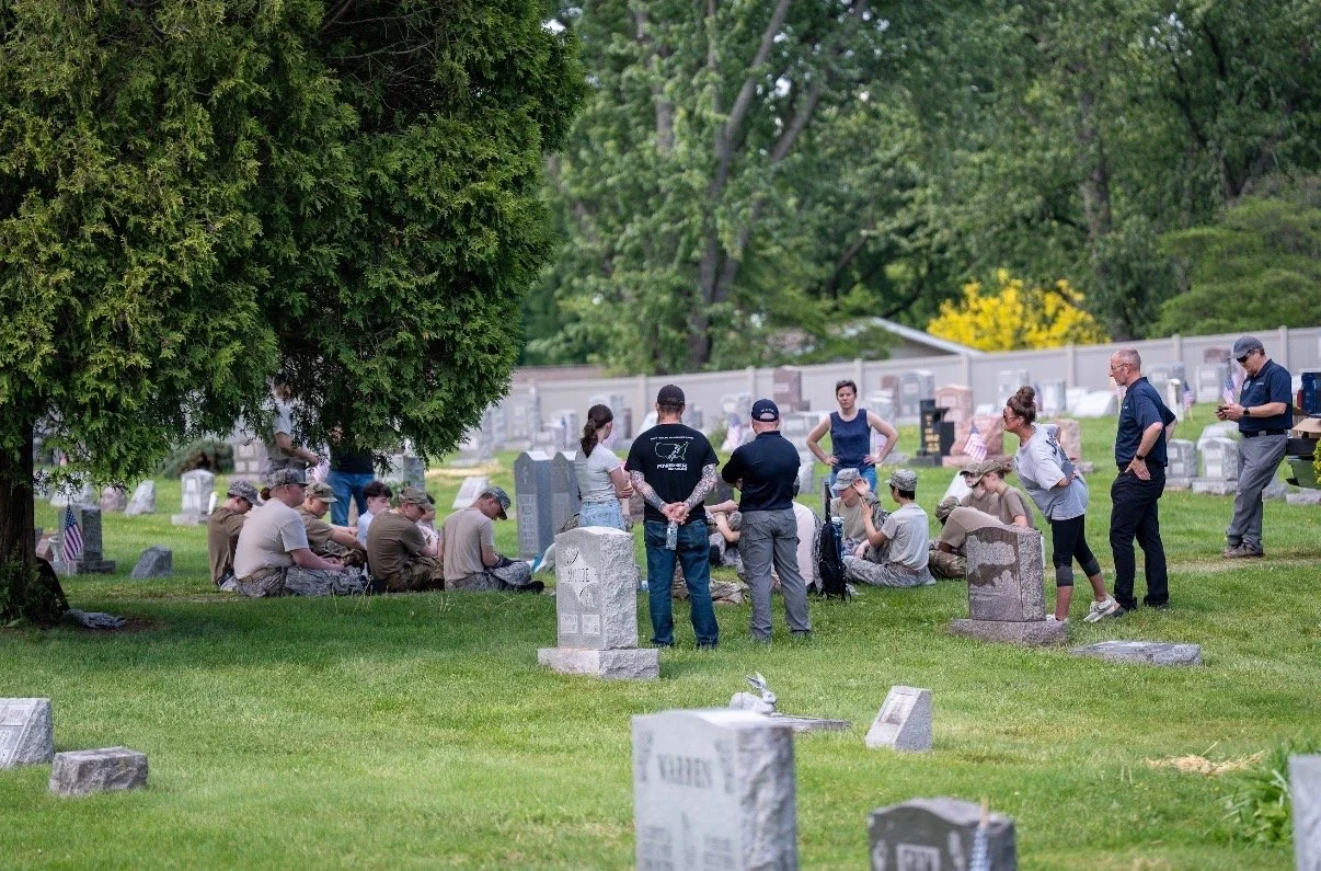 A group of people gathered in a cemetery, sitting and standing around a grave under a tree, with headstones and trees in the background.