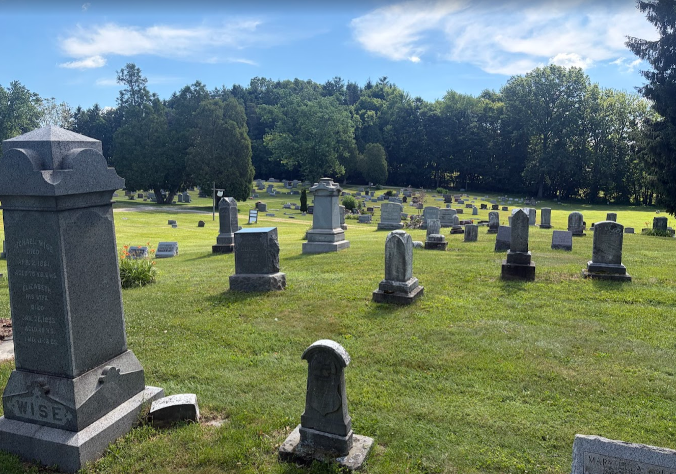 A cemetery with numerous gravestones on a green lawn under a partly cloudy blue sky, surrounded by trees.