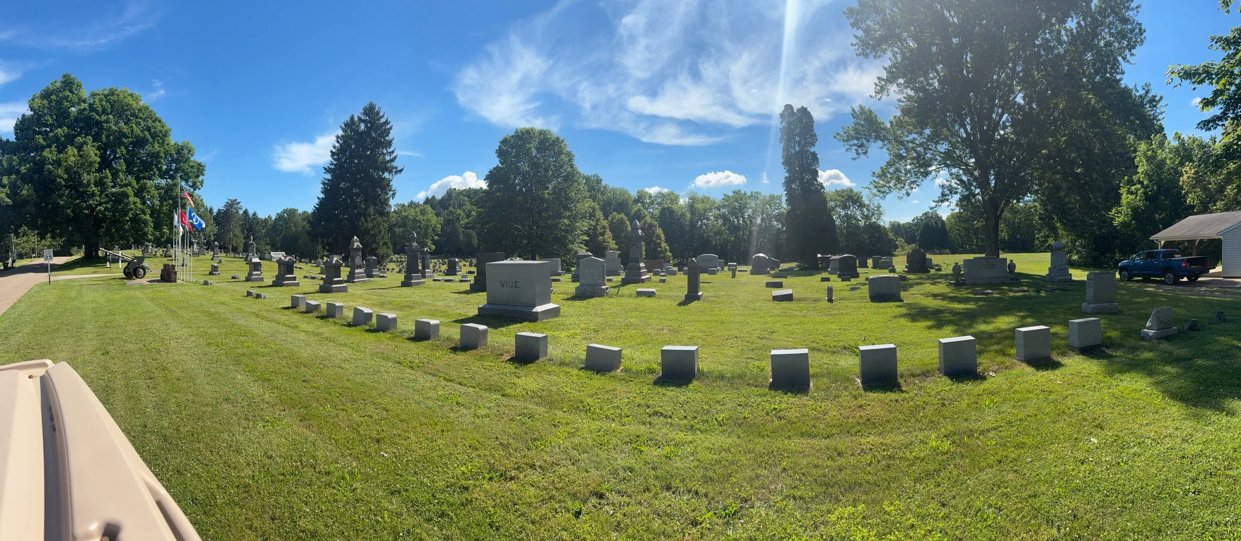 View of a cemetery with numerous gravestones and monuments, green grass, trees, a small building, and a car in the background on a sunny day with a bright blue sky and white clouds.