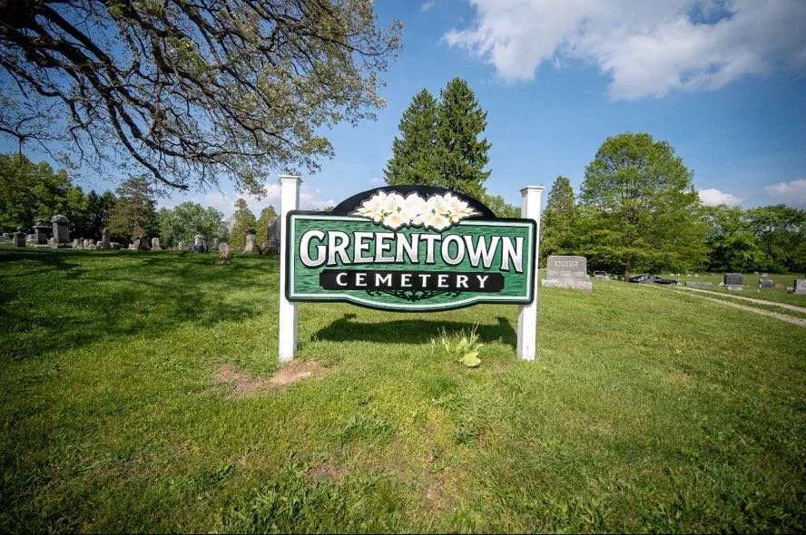 Sign for Greentown Cemetery with lush green grass, trees, and headstones in the background.