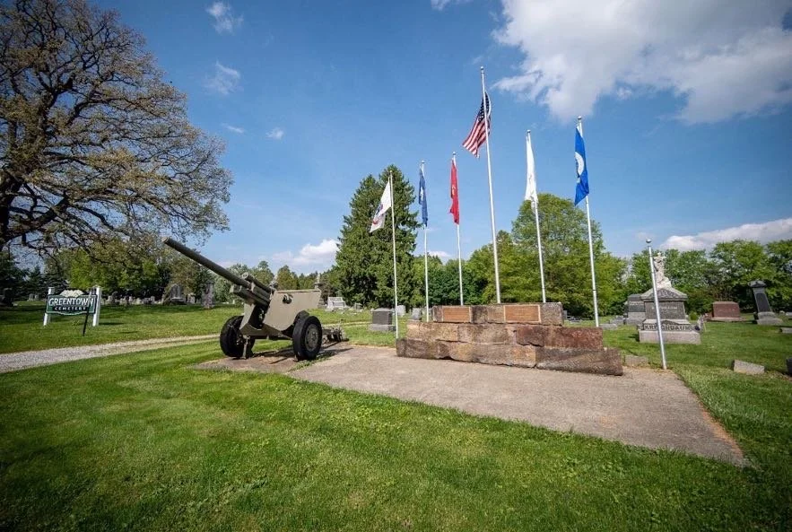 A display with six flags on poles, an artillery cannon, and several gravestones in a cemetery, with a sign reading 'Greentown Cemetery' in the background.