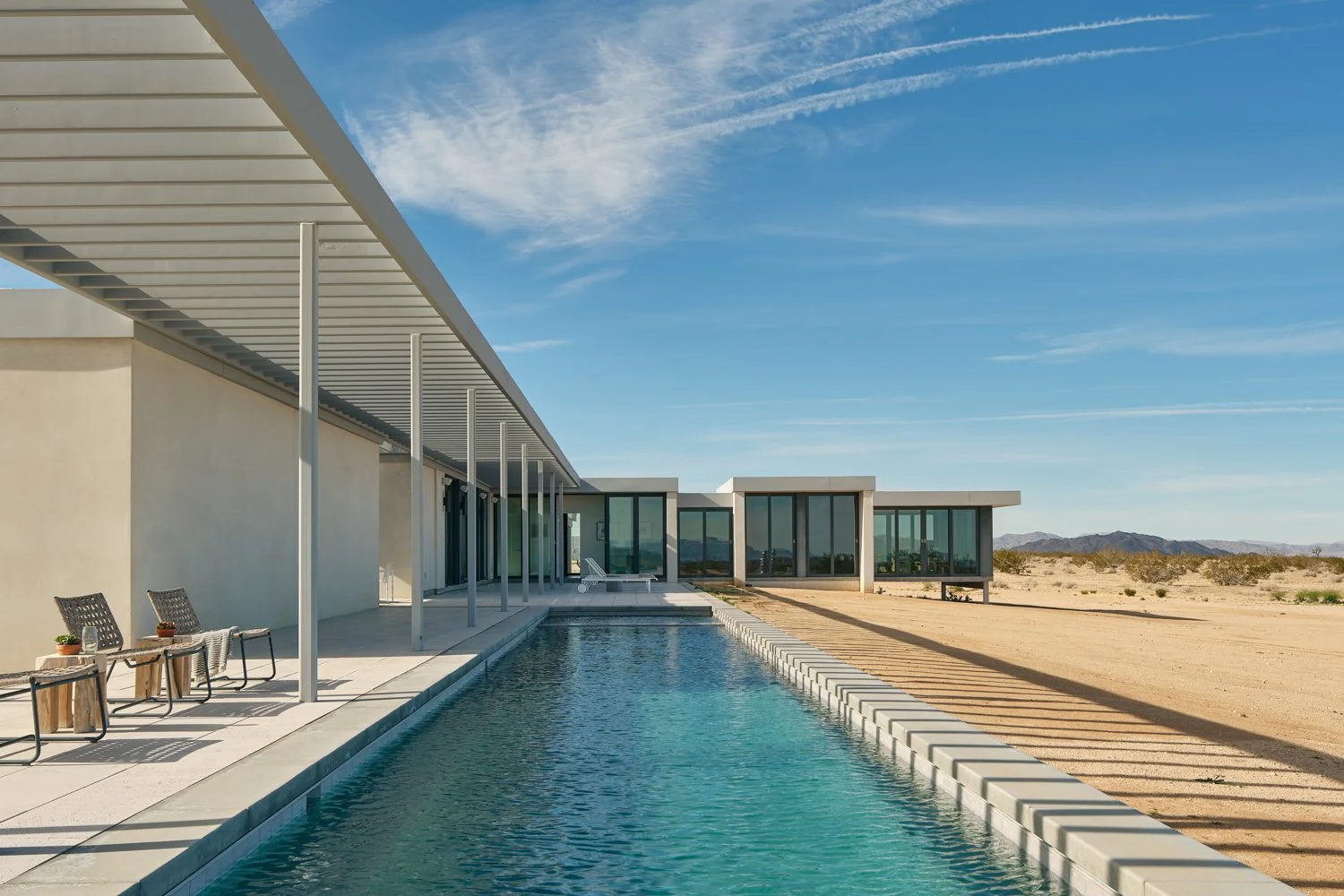 Modern house with flat roof and large windows, an outdoor pool, and desert landscape in the background under a blue sky.